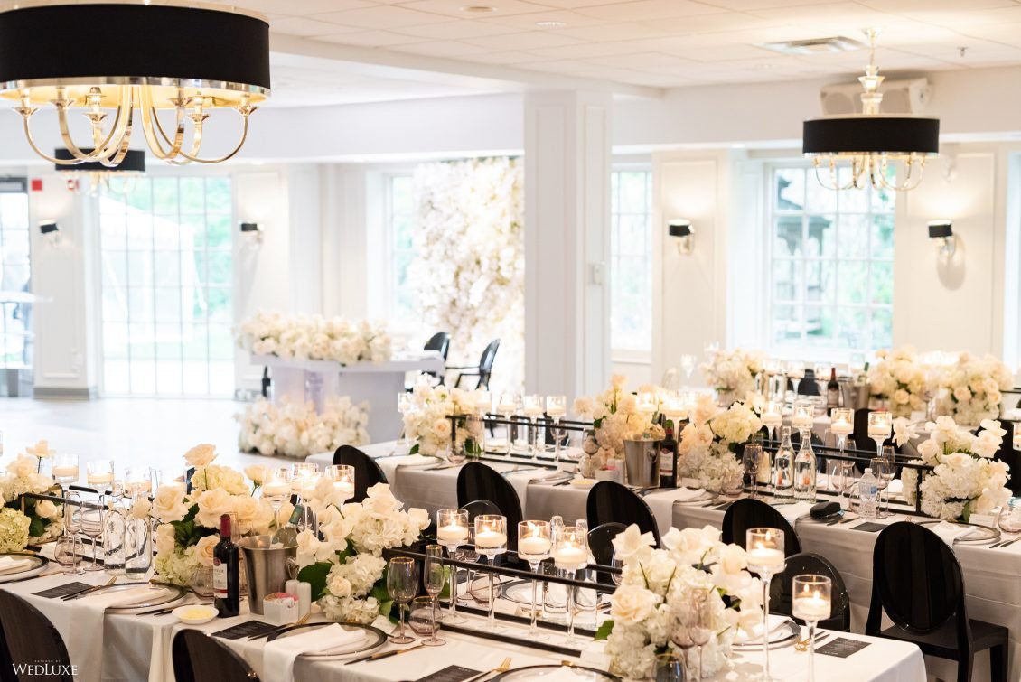 Elegant banquet hall decorated for a formal event with long tables covered in white tablecloths, adorned with white flower arrangements, lit candles, wine bottles, and glassware. Black chairs surround the tables, and the room has large windows lettin