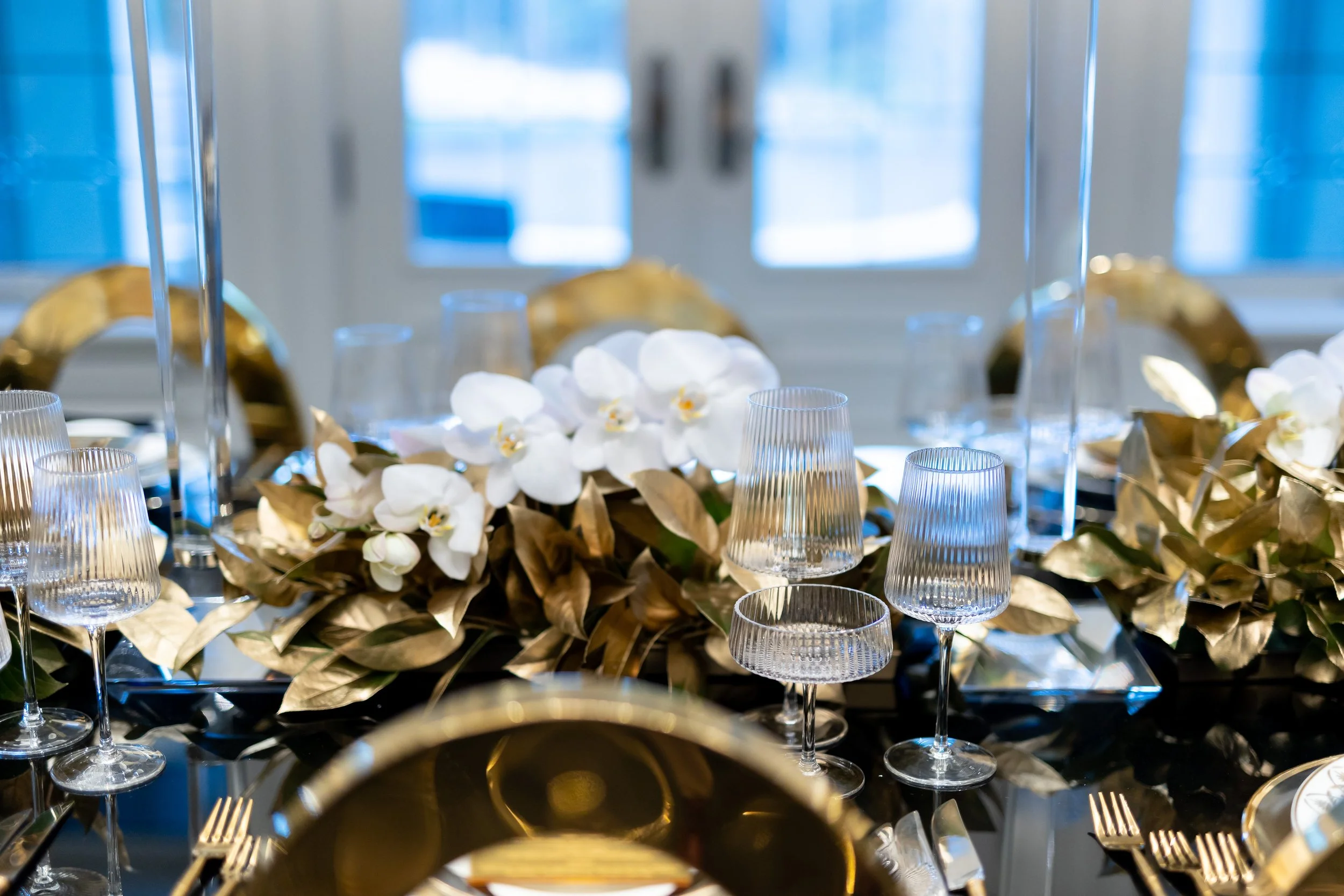 Elegant dining table with white orchids, gold leaves, and glassware, set for a formal event.