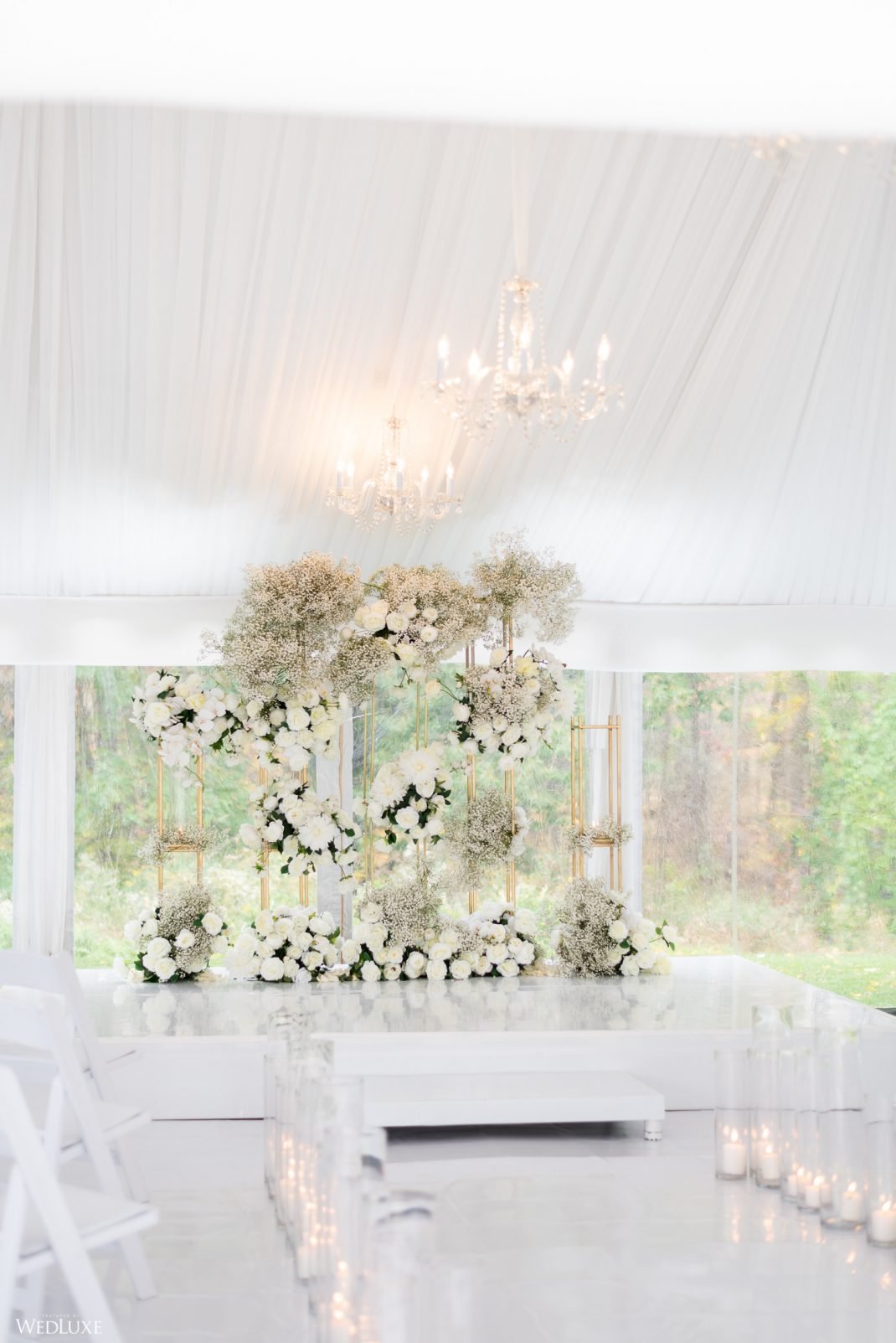Elegant wedding ceremony setup with white floral arrangements, gold accents, and chandeliers in a tented venue with natural light.