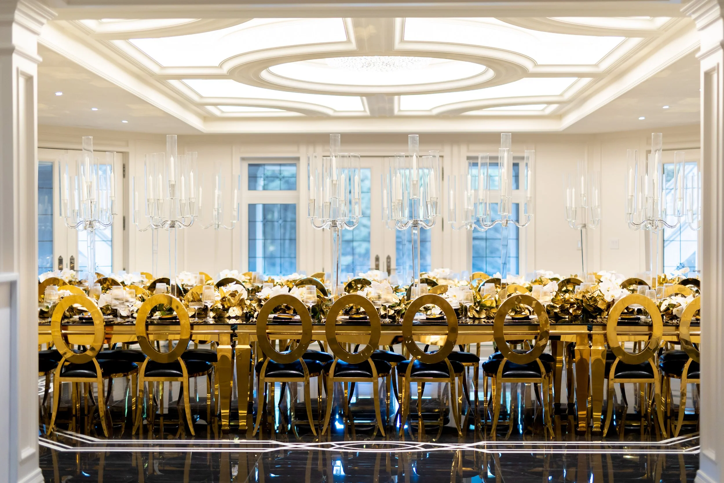 Elegant banquet hall with a long gold and black dining table, decorated with white flowers, gold chairs, and tall glass candle holders, illuminated by a large ornate chandelier and natural light from windows.