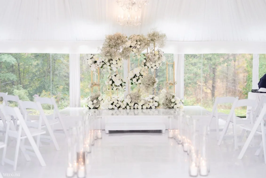 Elegant wedding ceremony setup with white chairs, floral arrangements, candles along the aisle, and a floral backdrop inside a bright tent with windows showing a wooded outdoor scene.