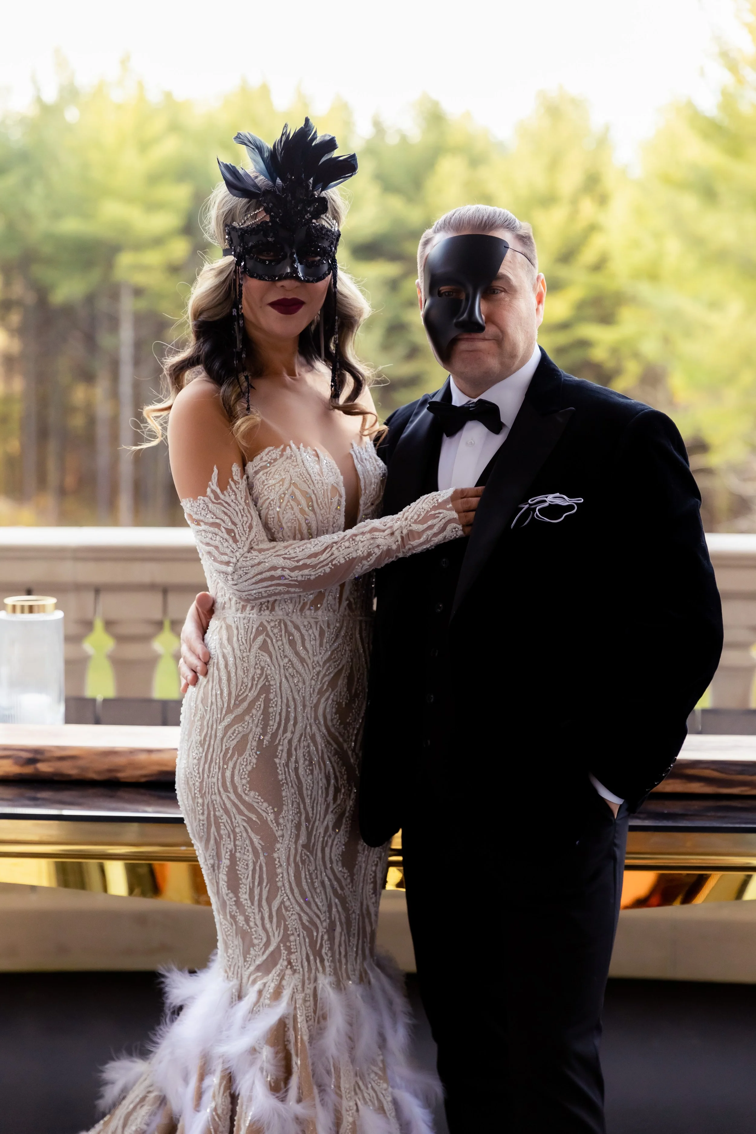 A woman in a lacy, feather-adorned wedding dress and a man in a tuxedo with a mask pose together at a wedding or formal event outdoors.