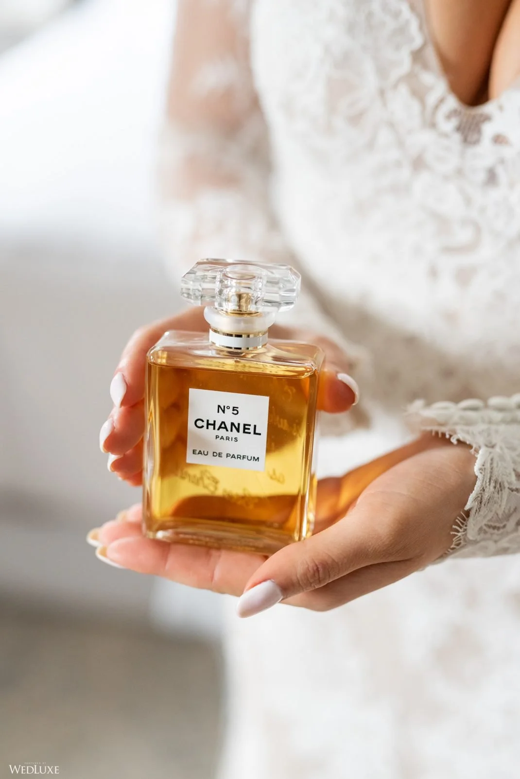 A woman holding a bottle of Chanel No. 5 perfume in both hands, wearing a white lace garment.