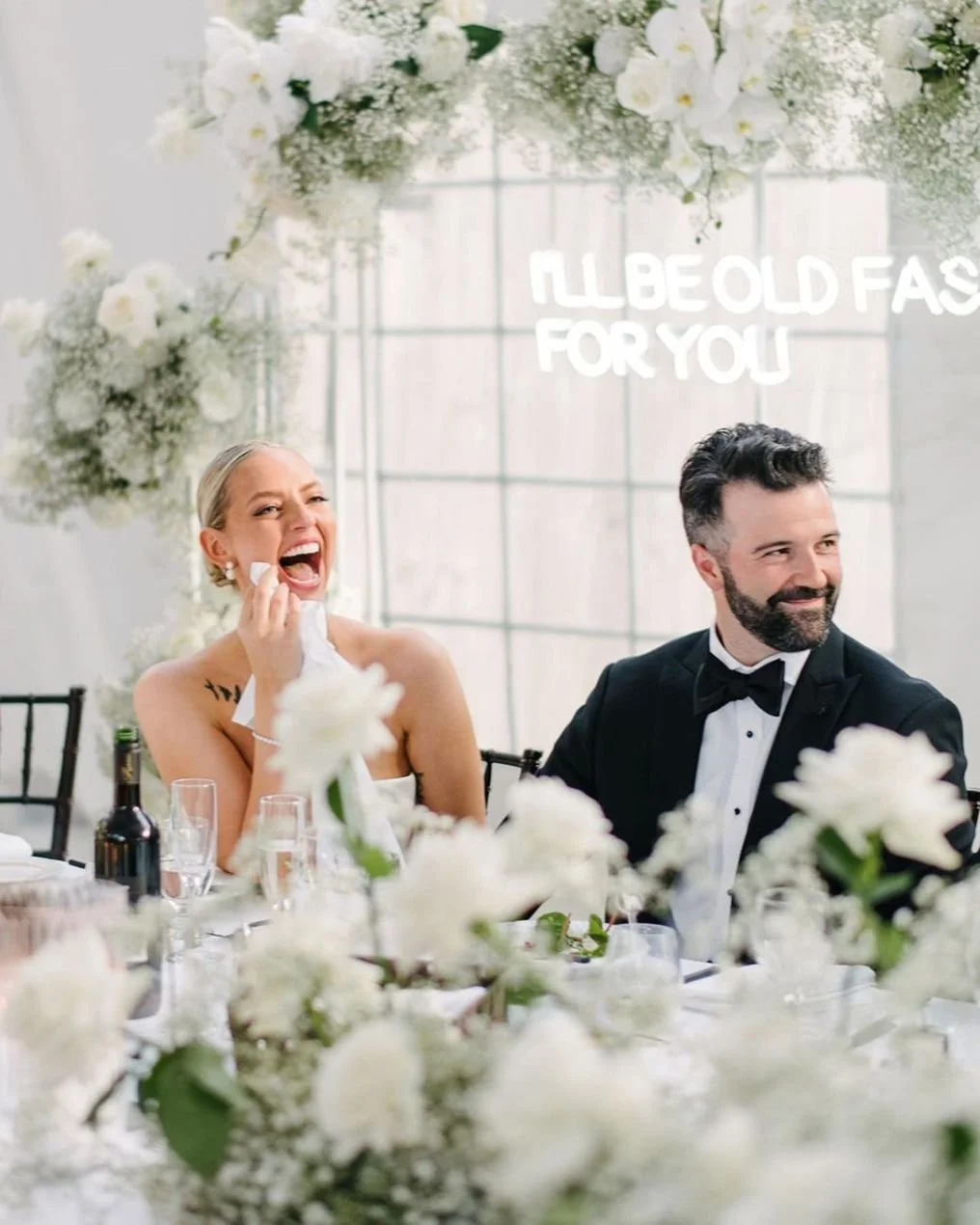 A woman and a man dressed in formal attire sit at a decorated table, smiling and laughing amidst white flowers, with a large window in the background and the words 'I'll be old fast for you' in white text.