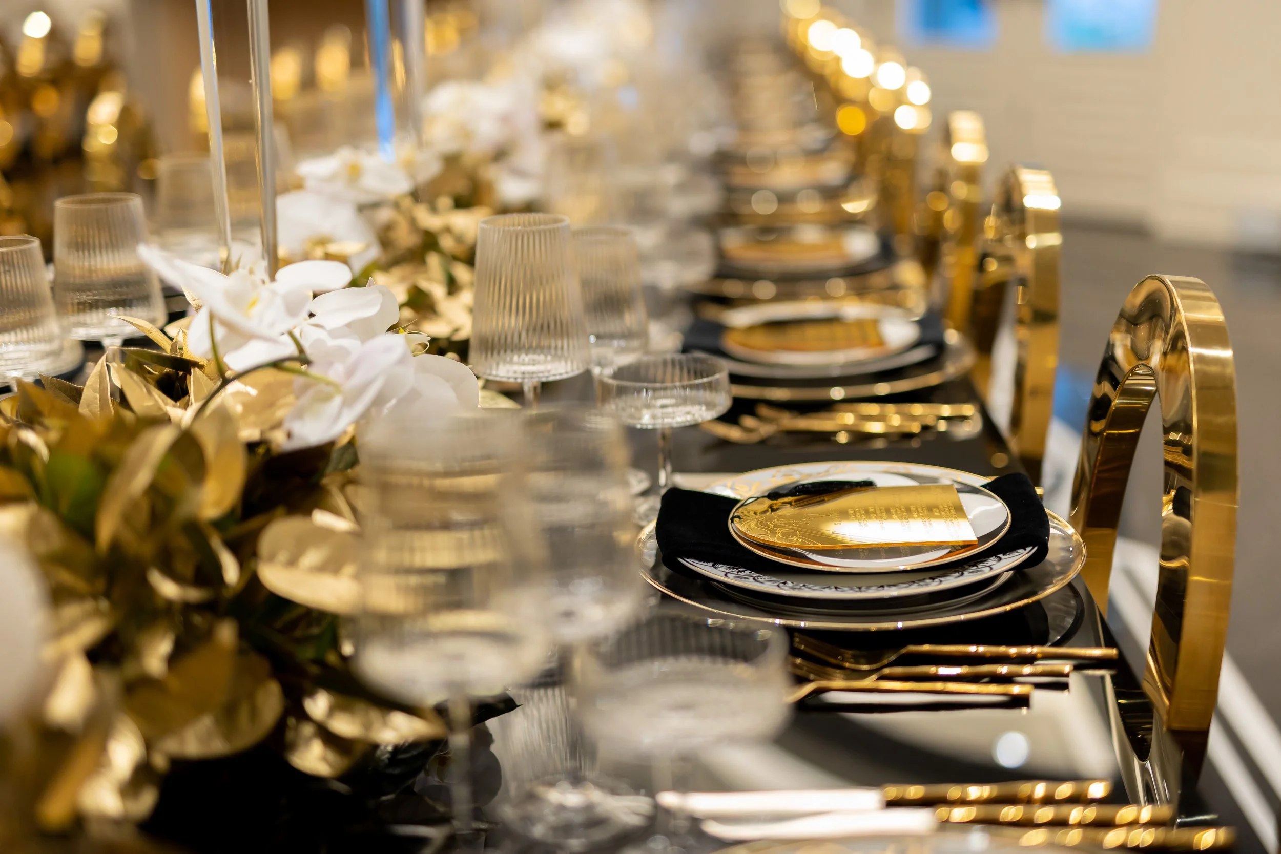 Elegant banquet table set with gold and black accents, white flowers, gold-rimmed plates, glassware, and black napkins.