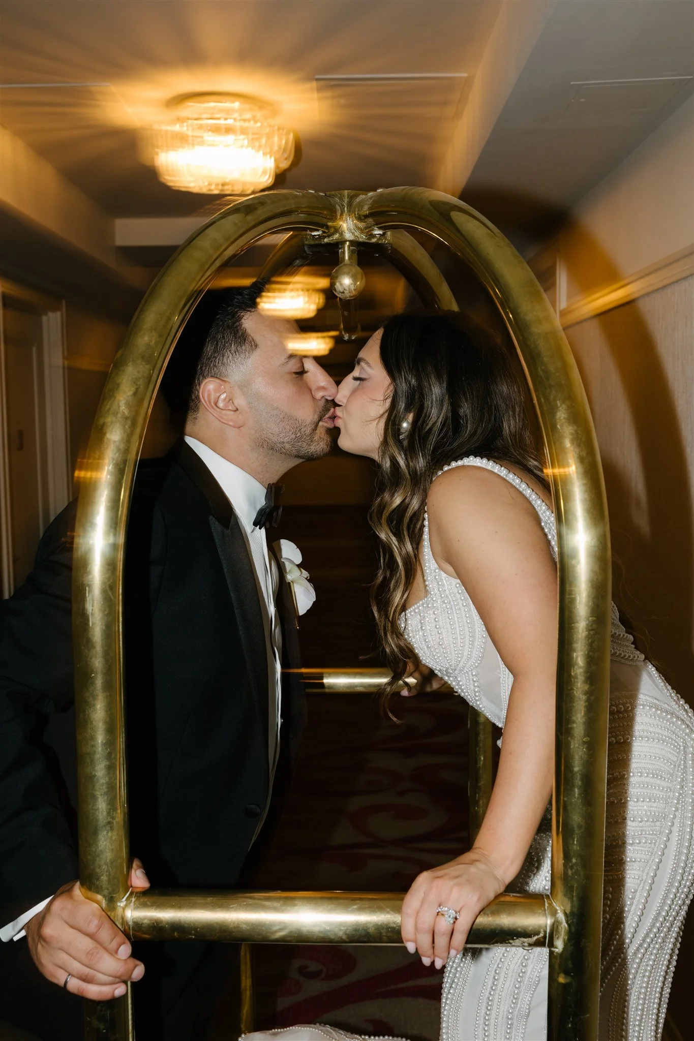 A groom and bride in wedding attire kissing through a golden luggage cart at their wedding reception.