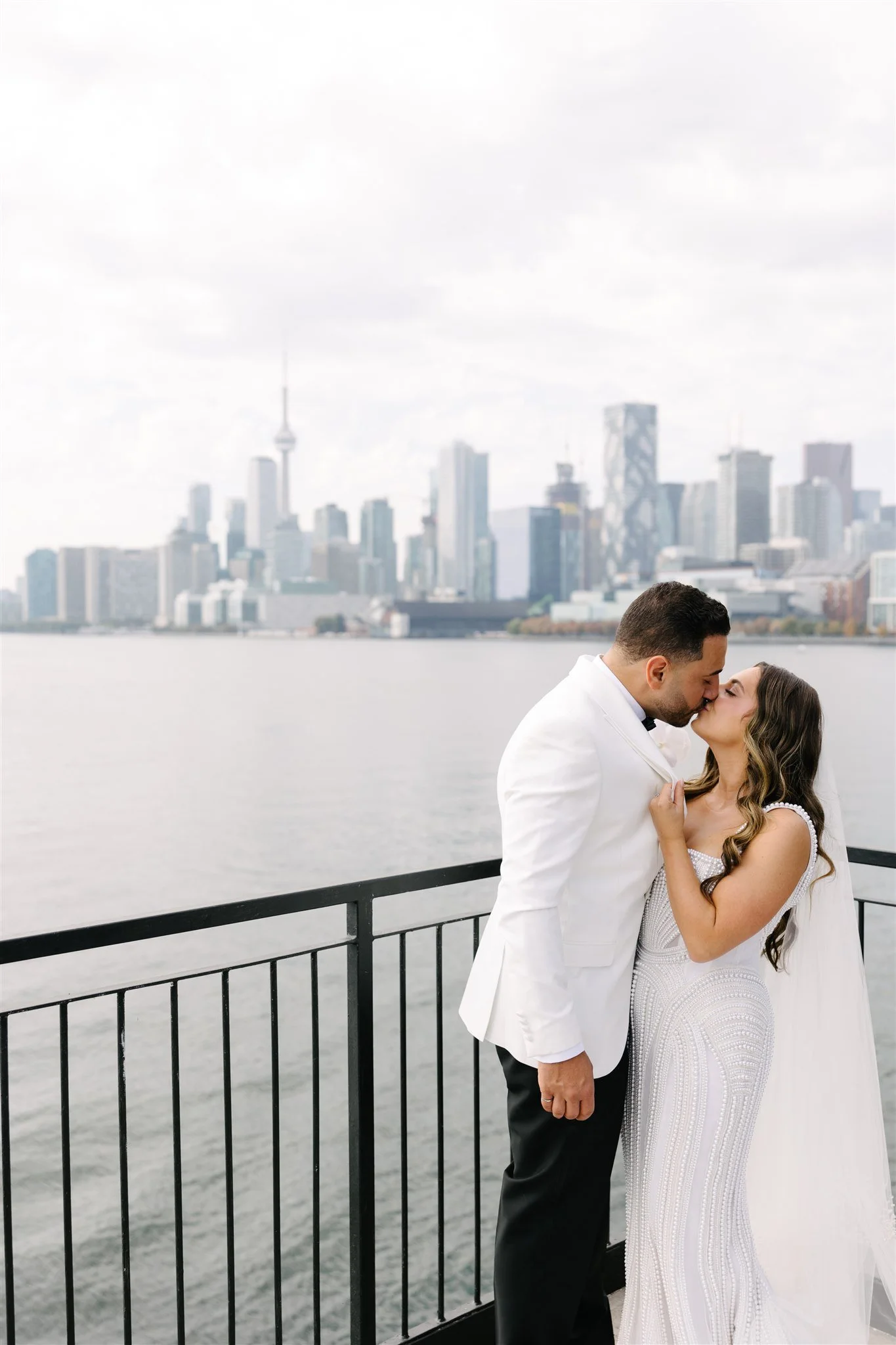 A bride and groom sharing a kiss on a balcony overlooking a city skyline with tall buildings and a body of water.