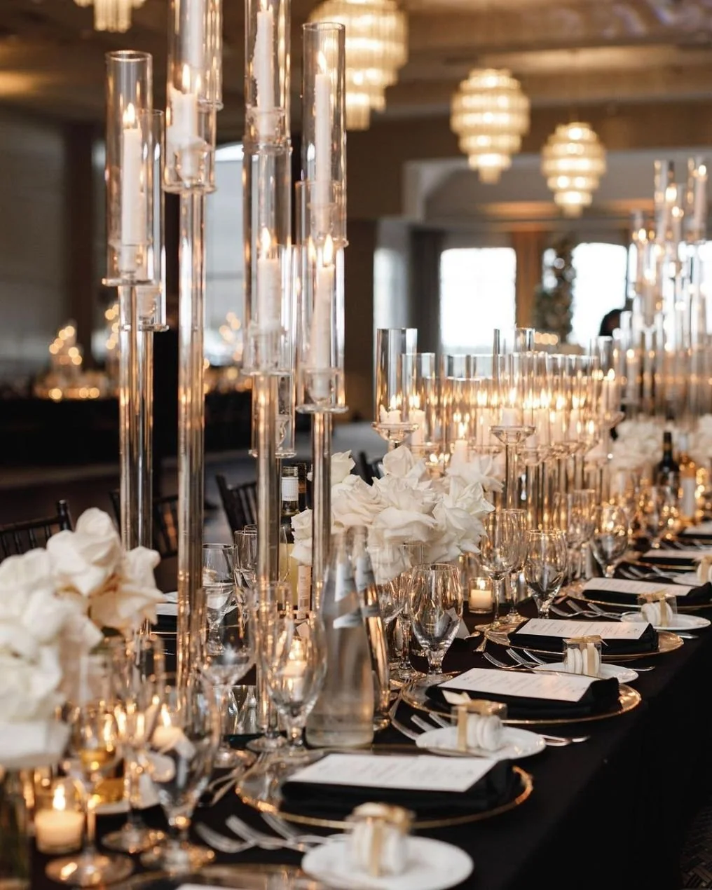 Elegant banquet table decorated with tall glass candleholders, white floral centerpieces, black napkins, champagne flutes, and white menus, with soft ambient lighting and chandeliers overhead.