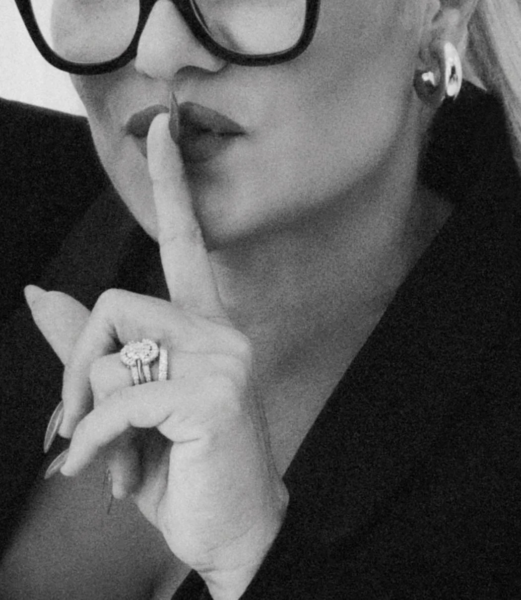 Close-up of a woman with glasses holding her finger to her lips regally, wearing jewelry, in black and white.