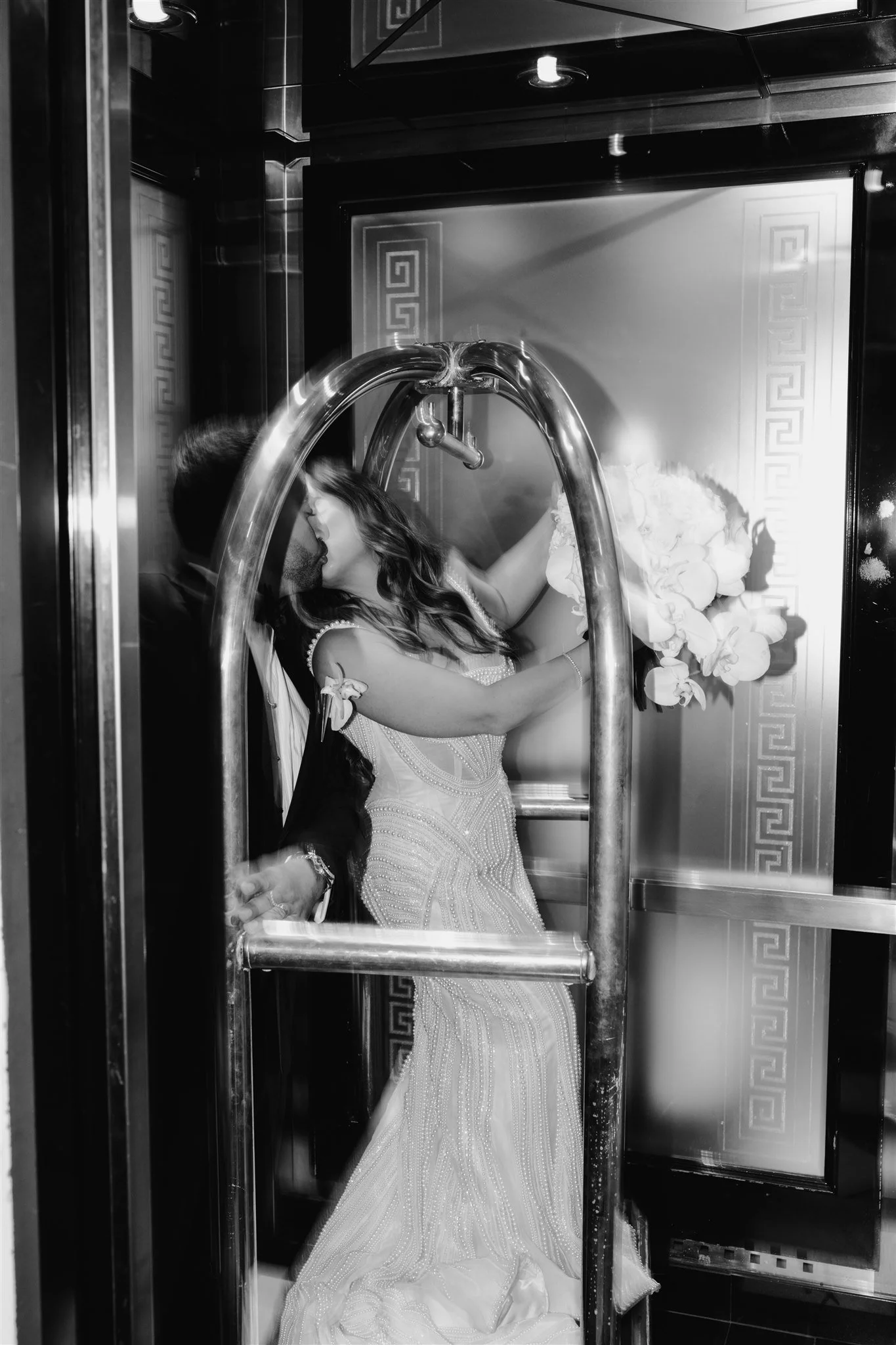 A couple kissing inside a glass elevator, with the woman holding a bouquet of flowers and the man in a suit.