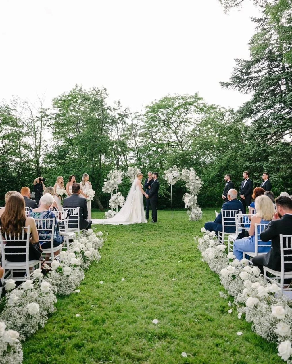 Outdoor wedding ceremony with bride and groom holding hands, officiant present, and bridal party standing nearby, surrounded by white floral decorations, seated guests, and lush green trees in the background.