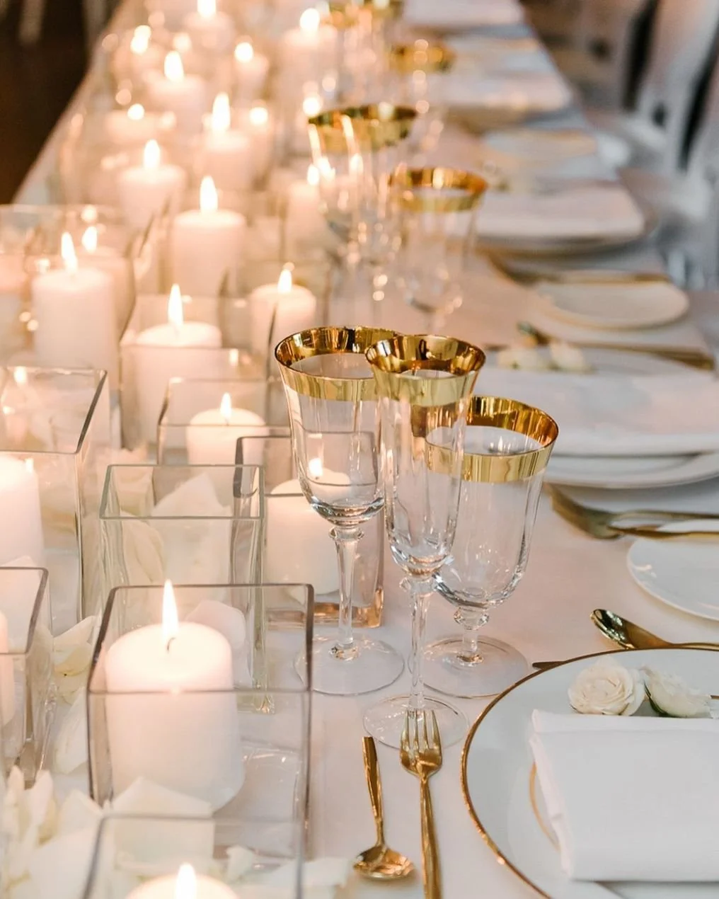 A formal dining table decorated with white candles in glass holders, crystal glasses with gold rims, and place settings with white plates, napkins, and gold utensils.