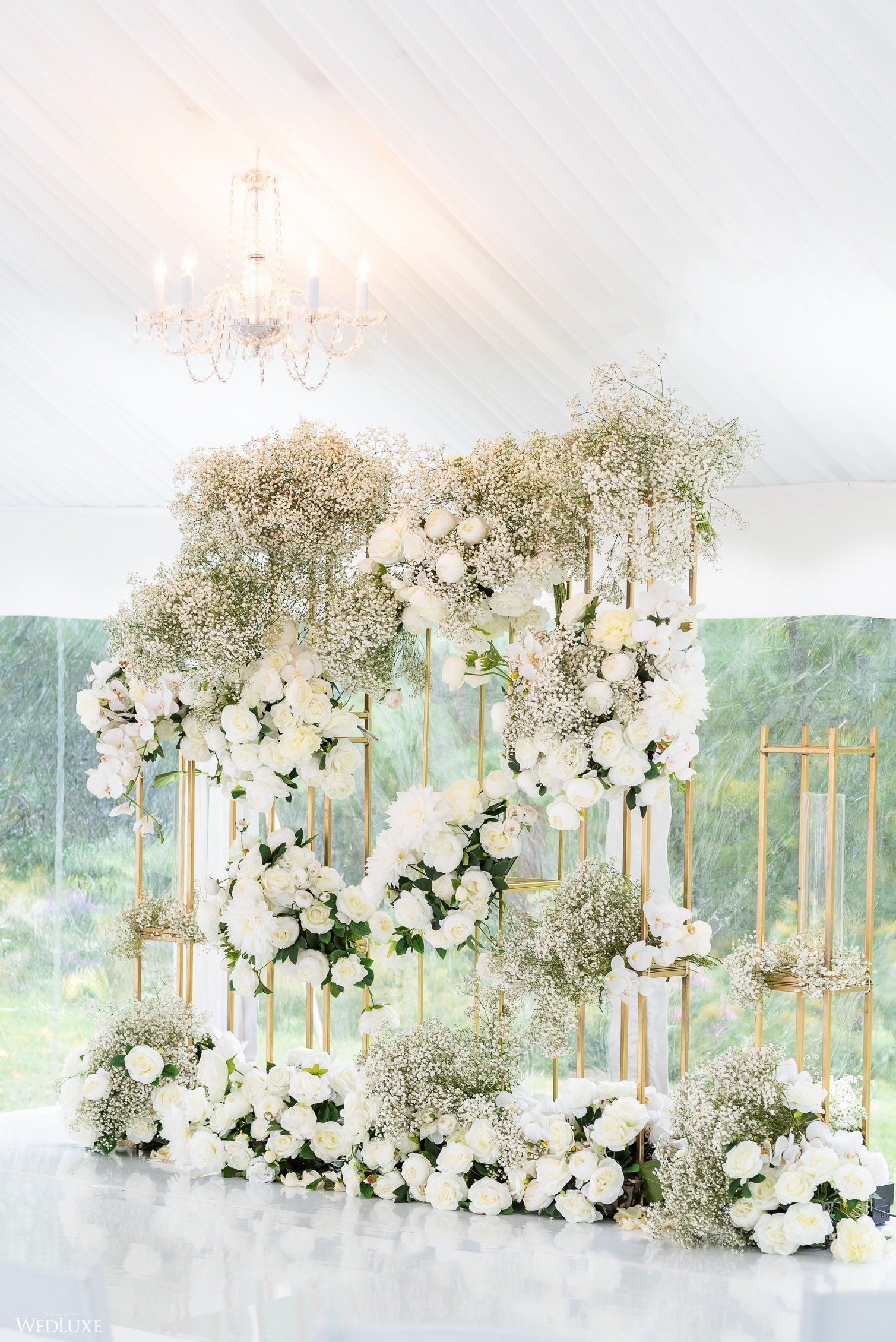 Elegant floral wedding backdrop with white flowers and golden frame, chandelier hanging from white ceiling, and a window with greenery outside.