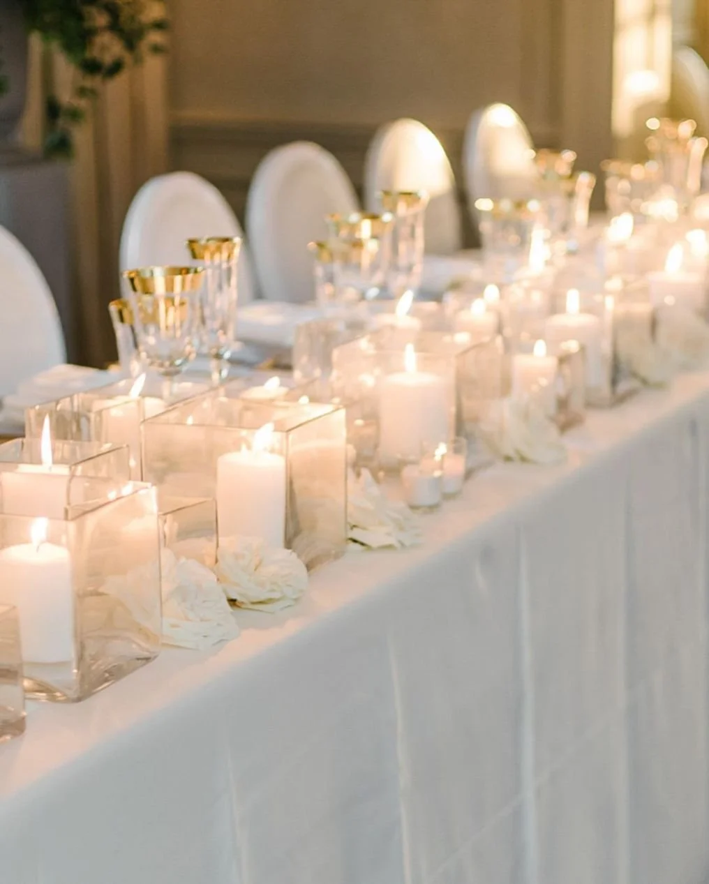 Elegant banquet table decorated with white candles in glass holders, white floral arrangements, and gold-rimmed glassware, set for a formal event.
