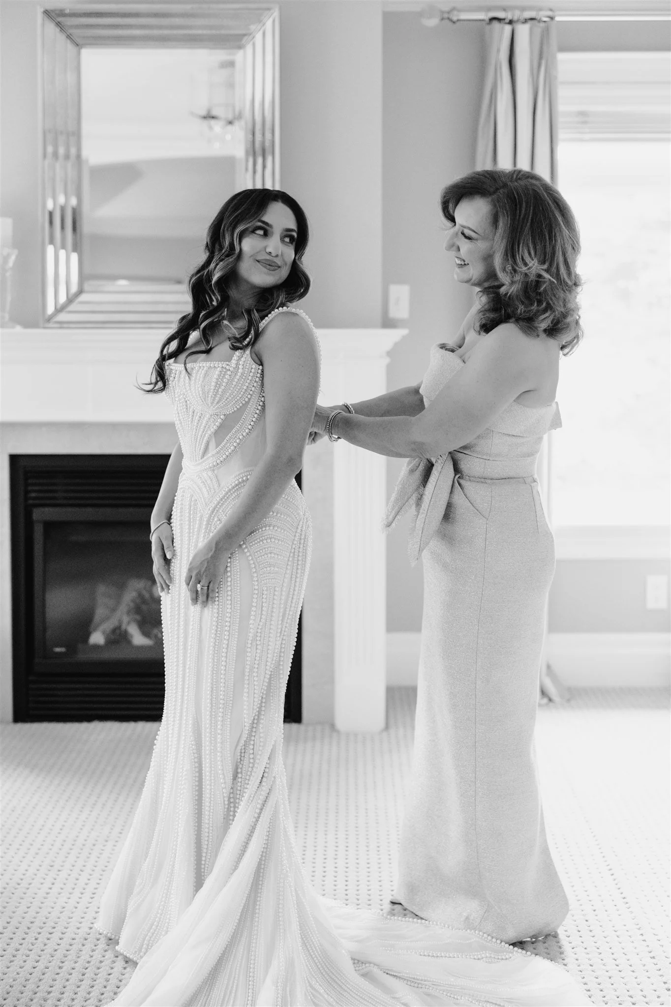 A bride is getting ready with help from a woman, possibly her mother, near a fireplace in a room with a large mirror and window. The bride is wearing a detailed wedding gown, and both women are smiling.