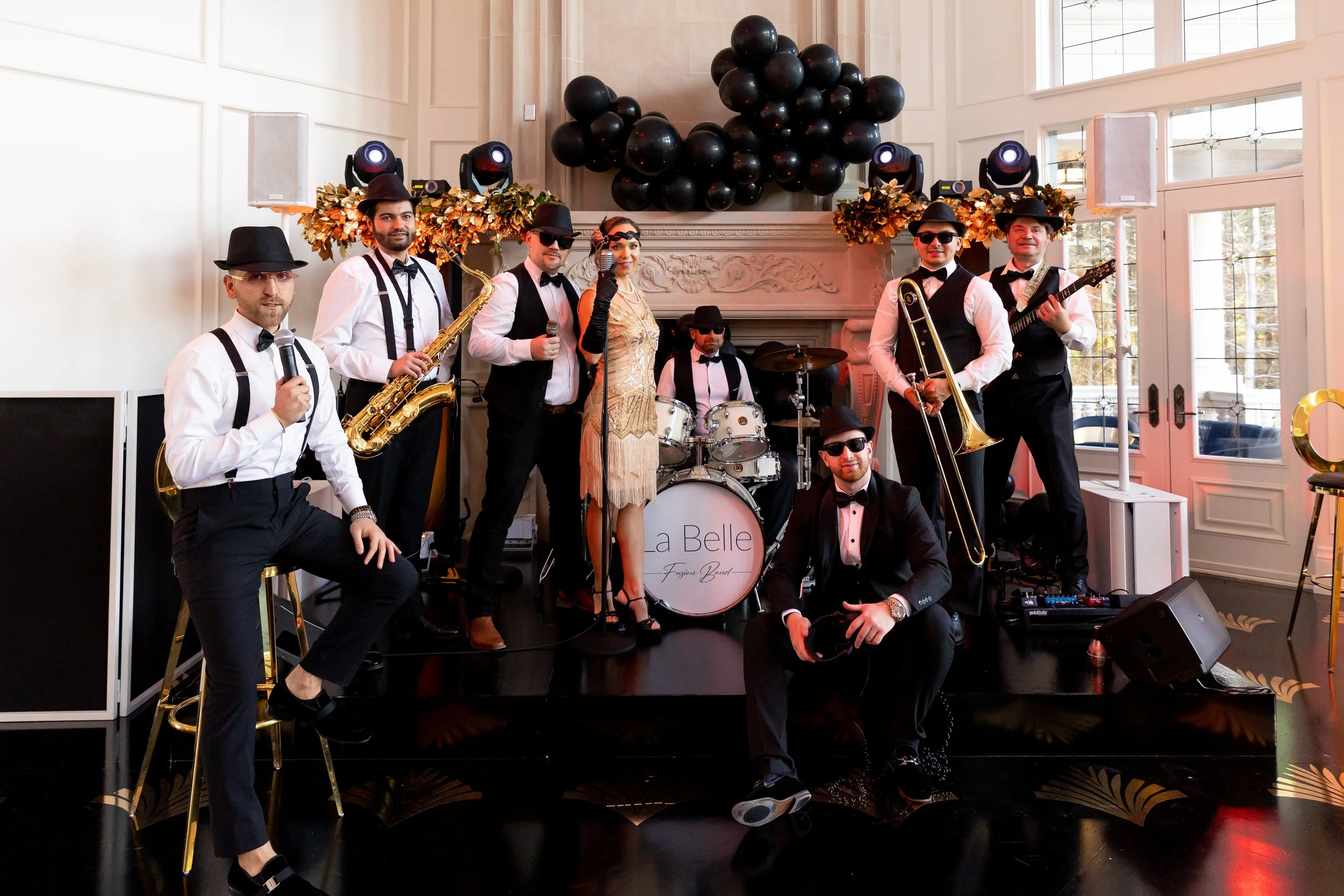 A band of nine musicians dressed in vintage style, posing with their instruments in a decorated indoor venue with black balloons and gold accents.