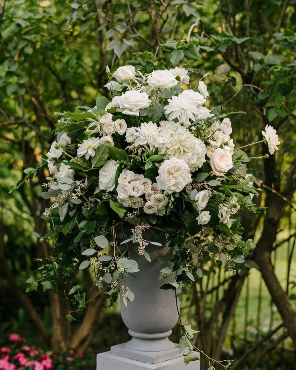 White flower arrangement in a gray urn on a pedestal outdoors, surrounded by greenery.