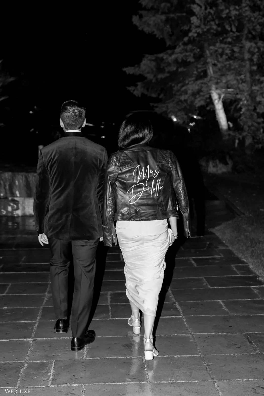A man and woman walking away on a paved path at night, the woman wearing a jacket with 'Mrs. Diddle' embroidered on the back.