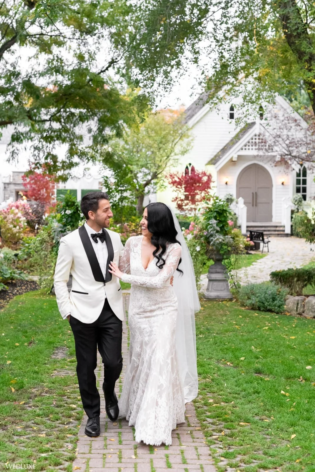 A newlywed couple walking arm in arm on a garden path, smiling at each other, with a white house and colorful trees in the background.