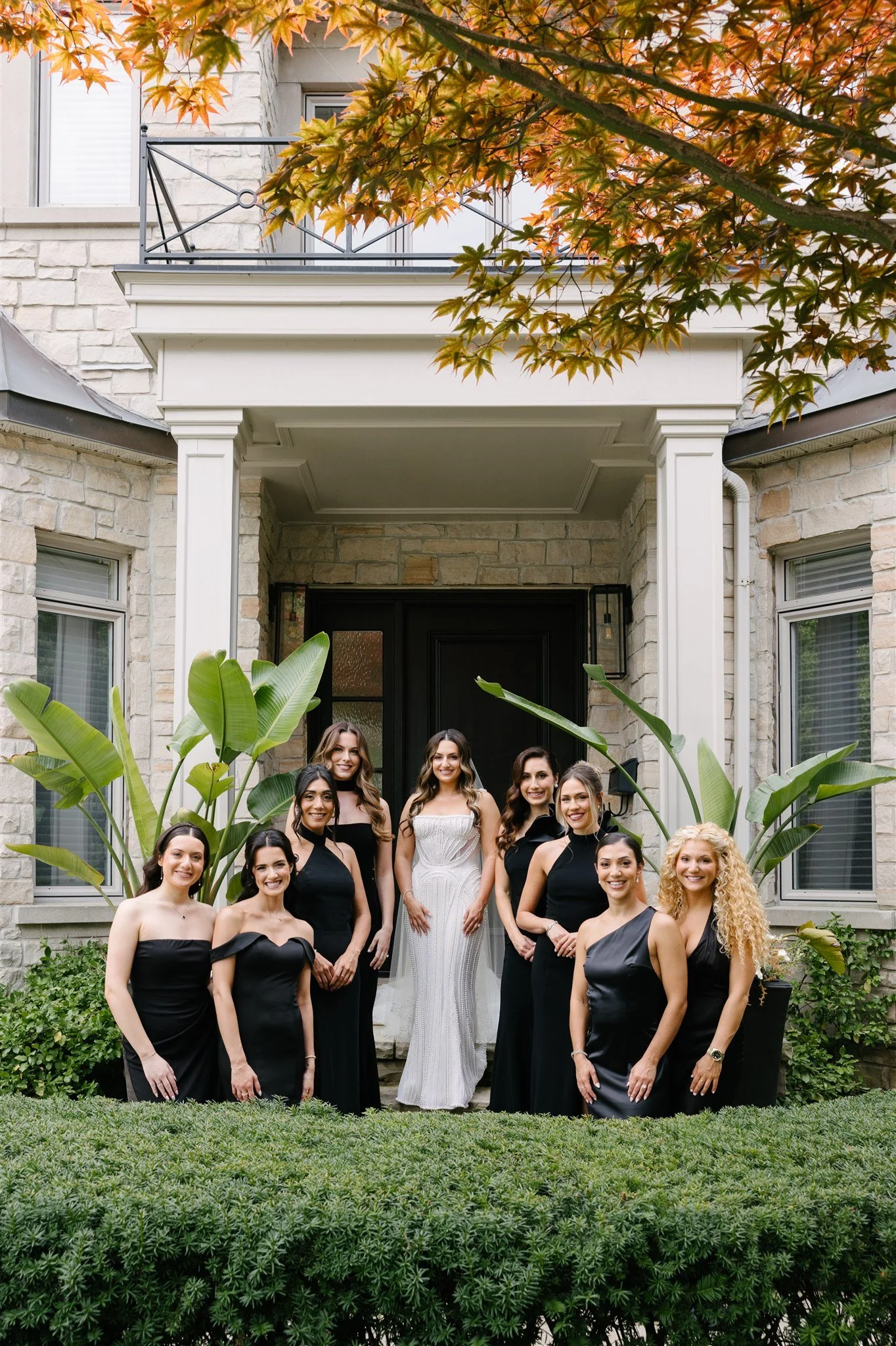 A group of ten women, dressed in formal black gowns with one in a white gown, standing on the steps of a stone house with large green plants in front and trees with orange leaves overhead.