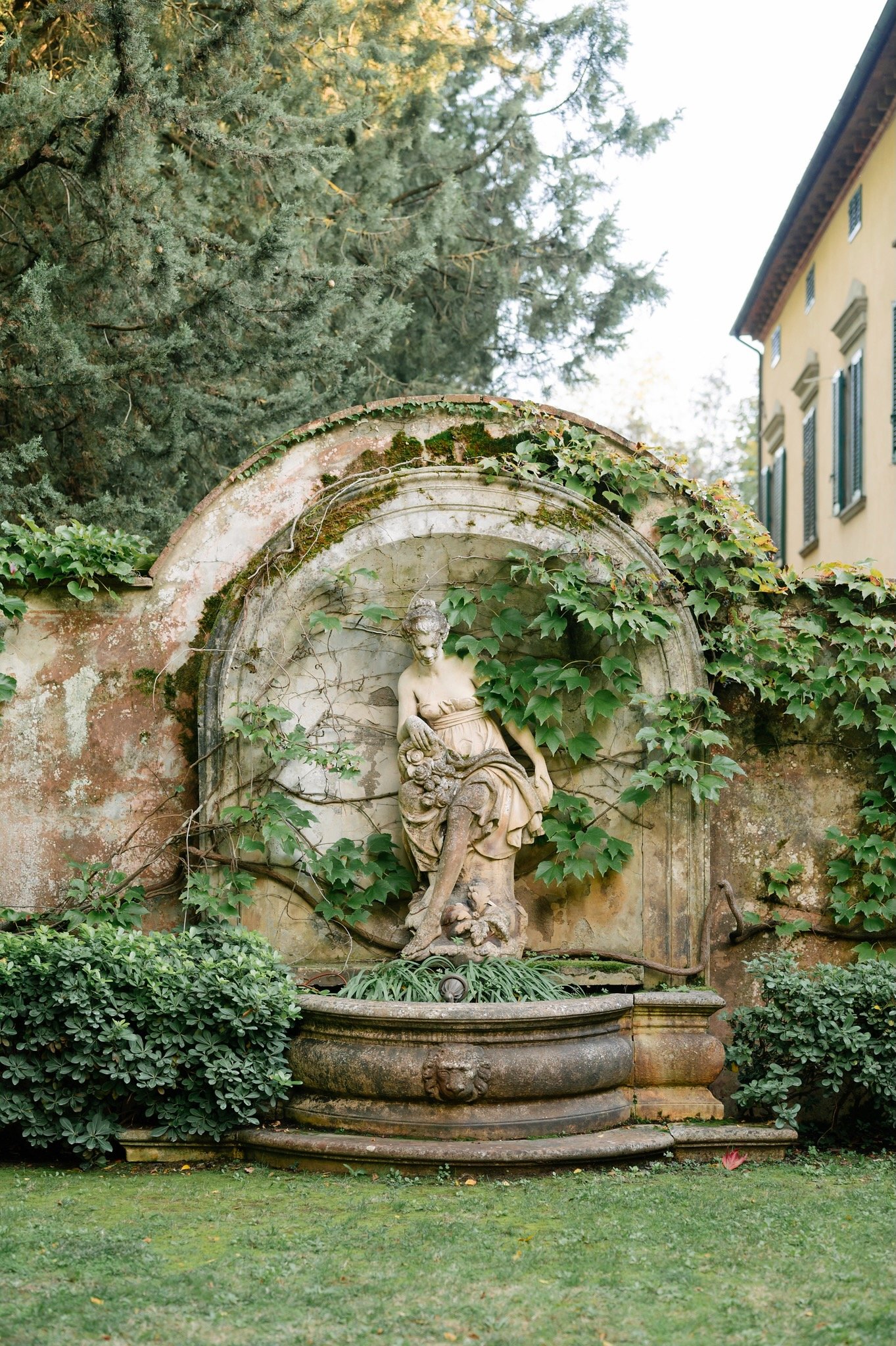 An old stone fountain featuring a classical statue of a woman, surrounded by green ivy and bushes, with trees and a building in the background.