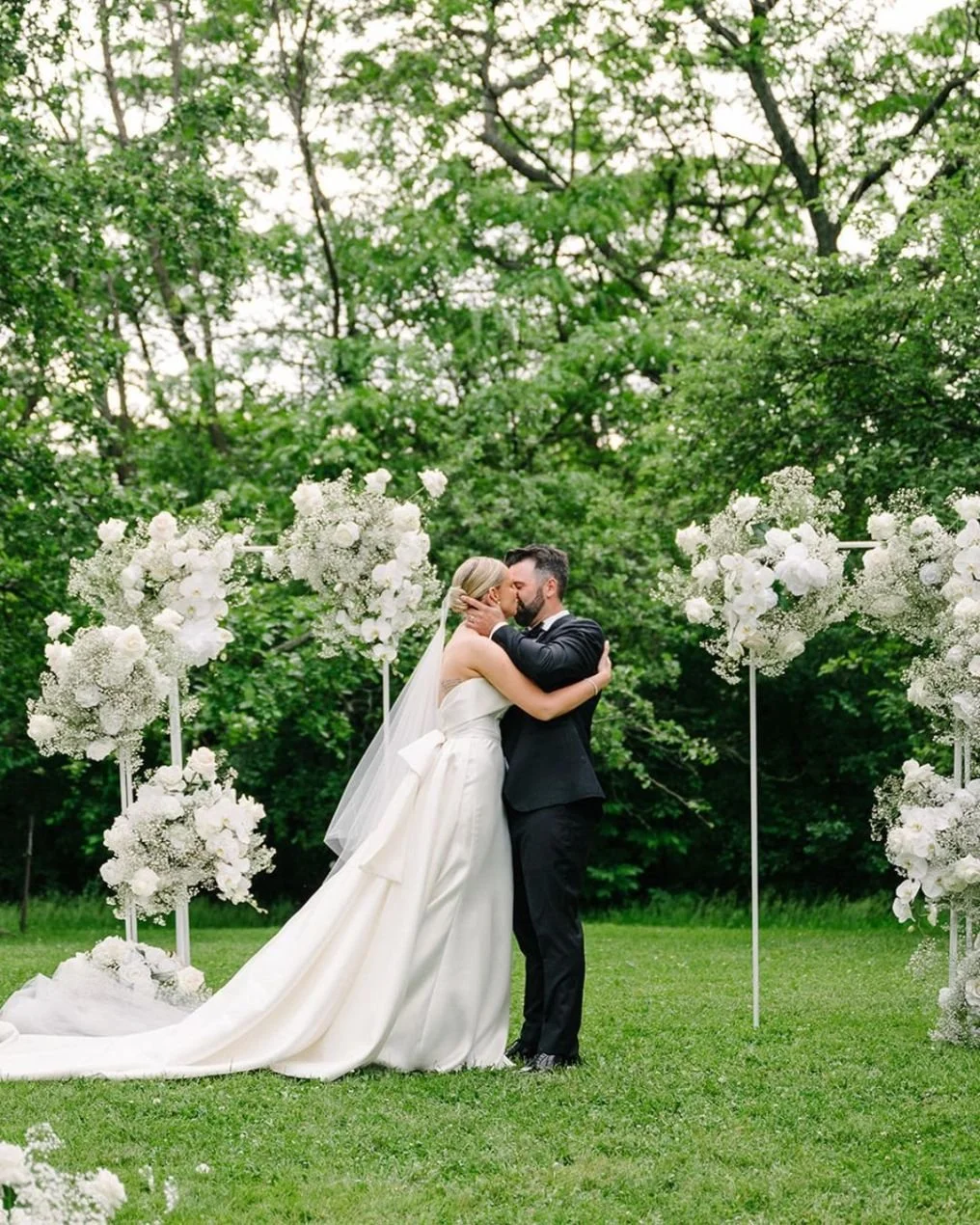 A bride and groom kiss during an outdoor wedding ceremony surrounded by white floral arrangements and greenery.