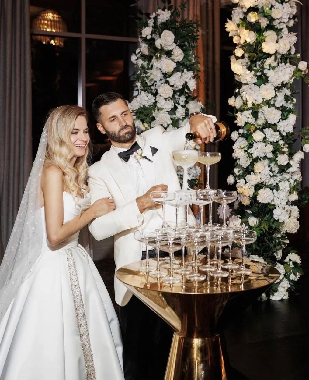 Bride and groom in wedding attire pouring champagne into a tower of glasses at their wedding reception, with large floral arrangements behind them.