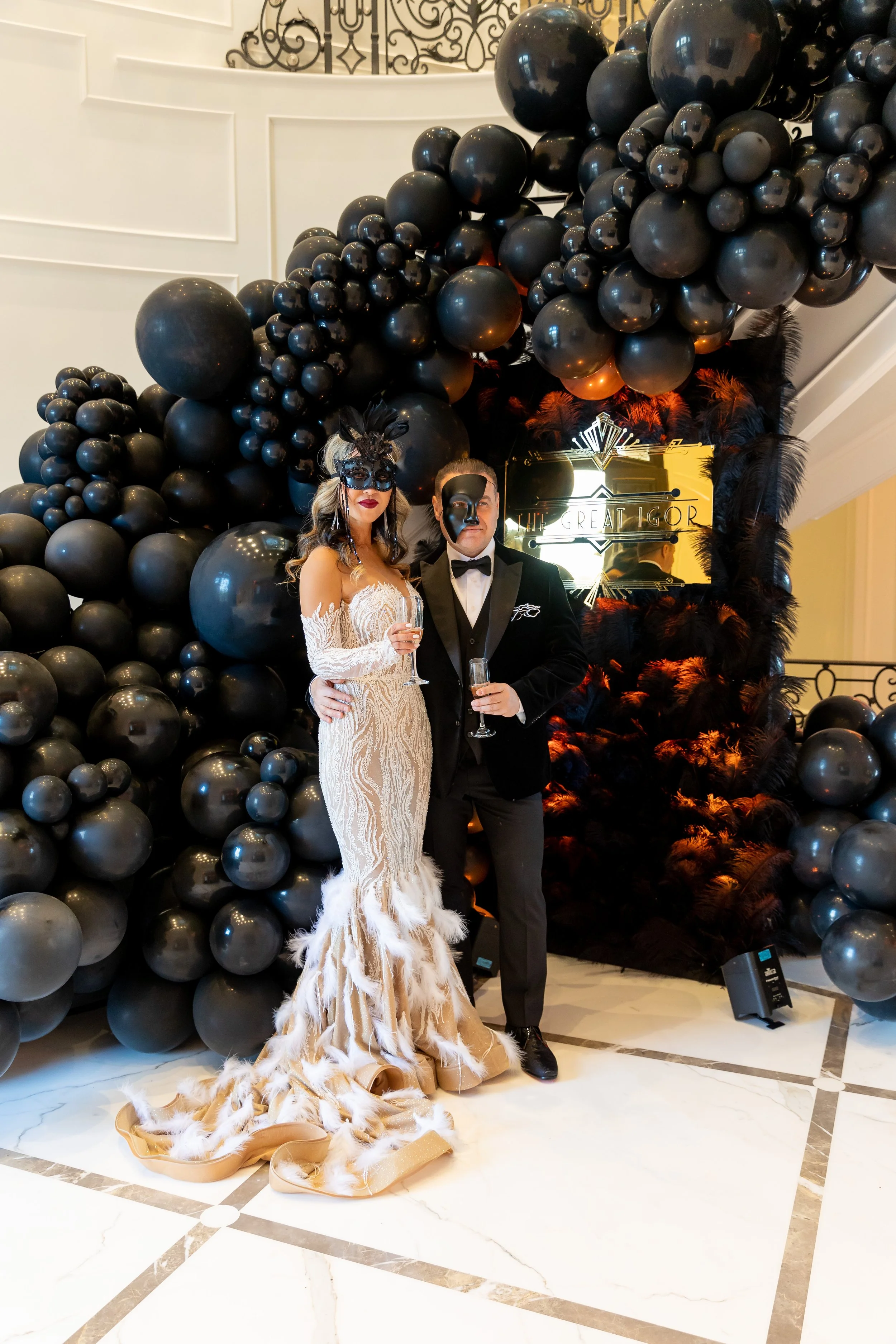 A woman and man in formal attire standing in front of a decorative black balloon arch and a black feathered backdrop with a sign that reads 'The Great Gigi.' The woman is wearing an ornate white gown with feathered hem and a black mask, and the man i