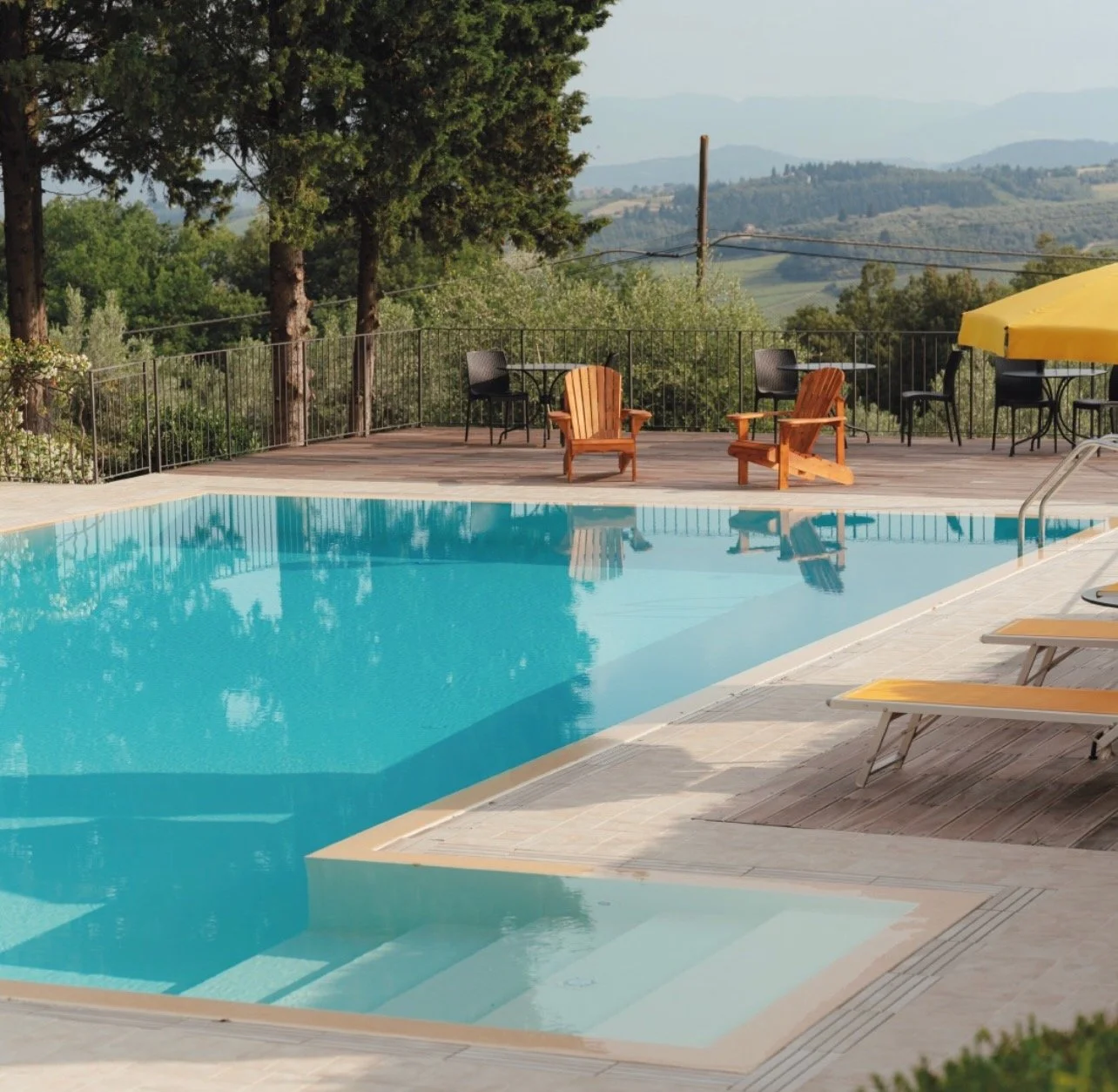 An outdoor swimming pool with a view of rolling hills and trees in the background. There are wooden chairs, Adirondack chairs, and black tables with chairs around the pool area, which is fenced with metal railings. A yellow umbrella is also visible.