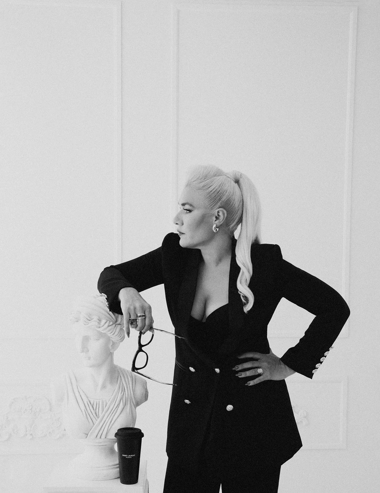 A woman with platinum blonde hair in a ponytail, dressed in a black blazer and trousers, holding glasses near a classical bust sculpture, in a monochrome setting.