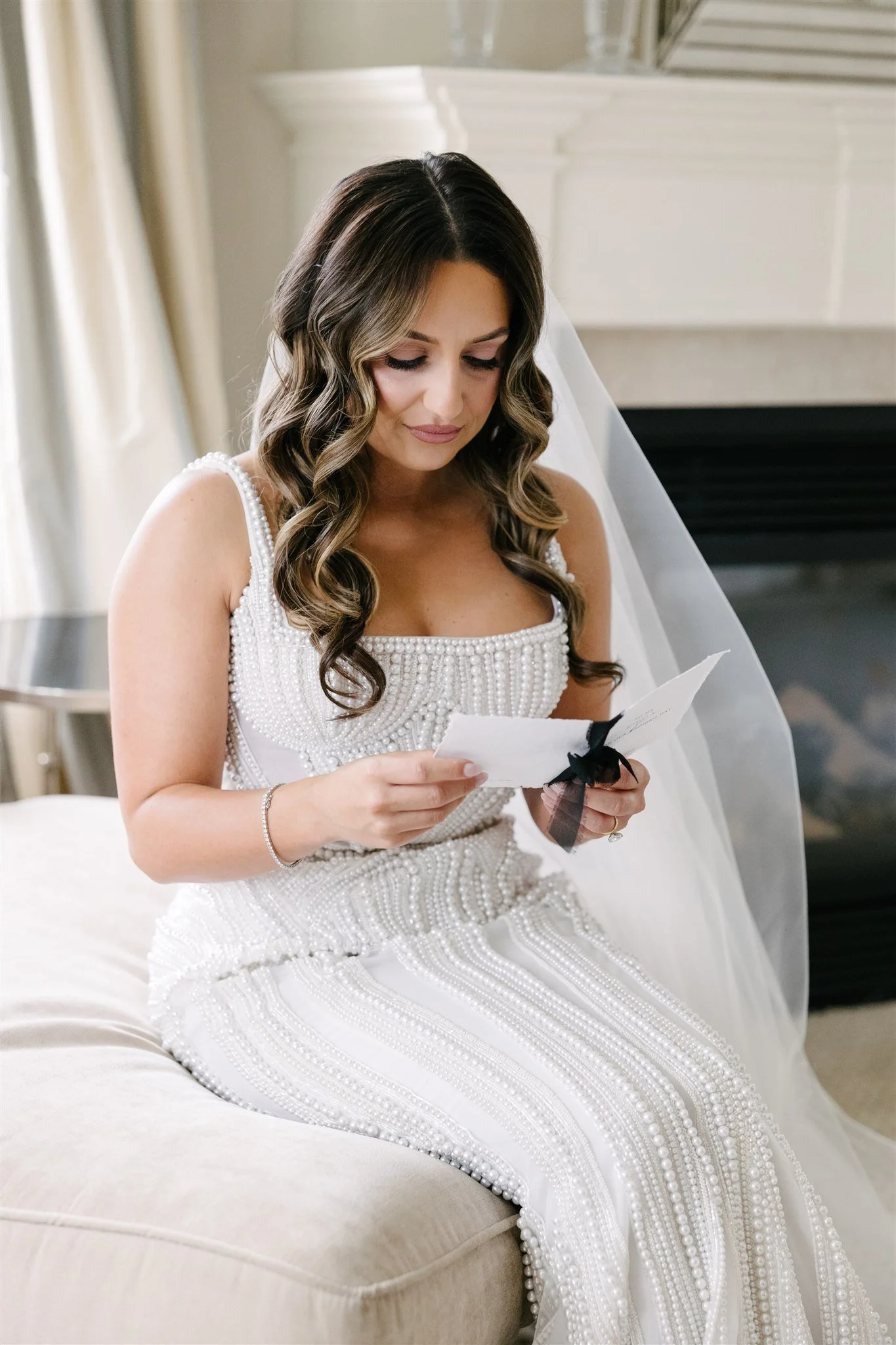 A woman in a white wedding dress with pearl embellishments is sitting on a beige couch, reading a card with a black ribbon.