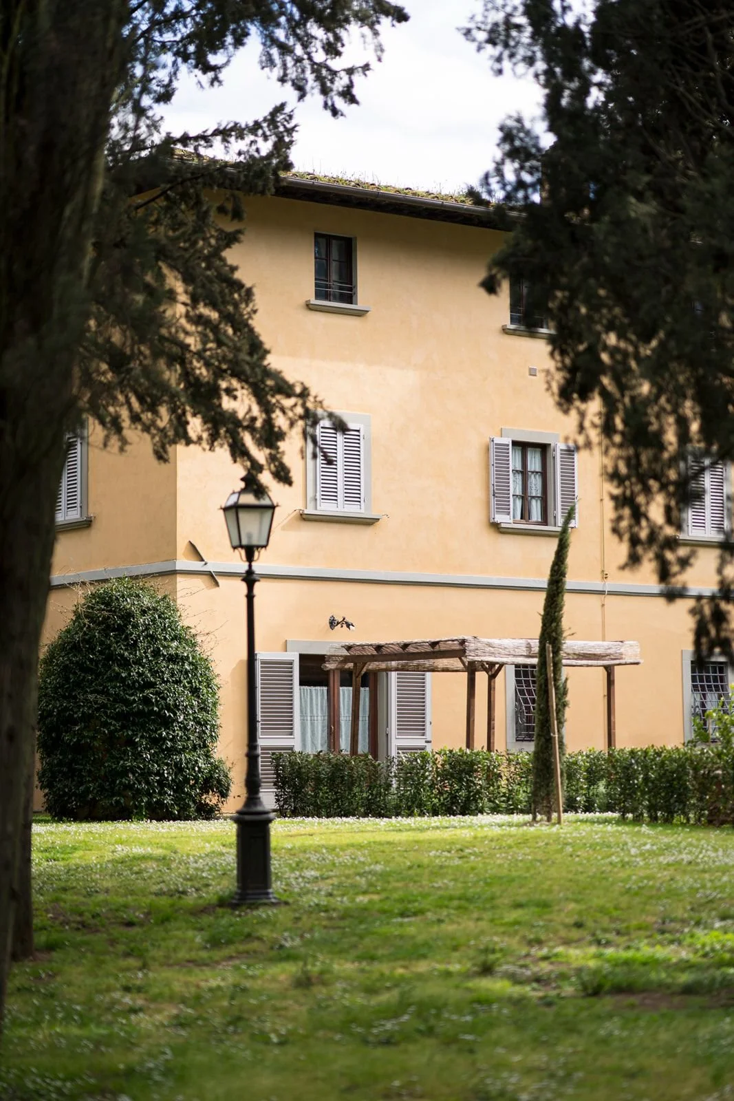 A yellow multi-story building with open window shutters, a small balcony, and a pergola in front, surrounded by green trees and bushes, with a grassy lawn and a lamppost in the foreground.