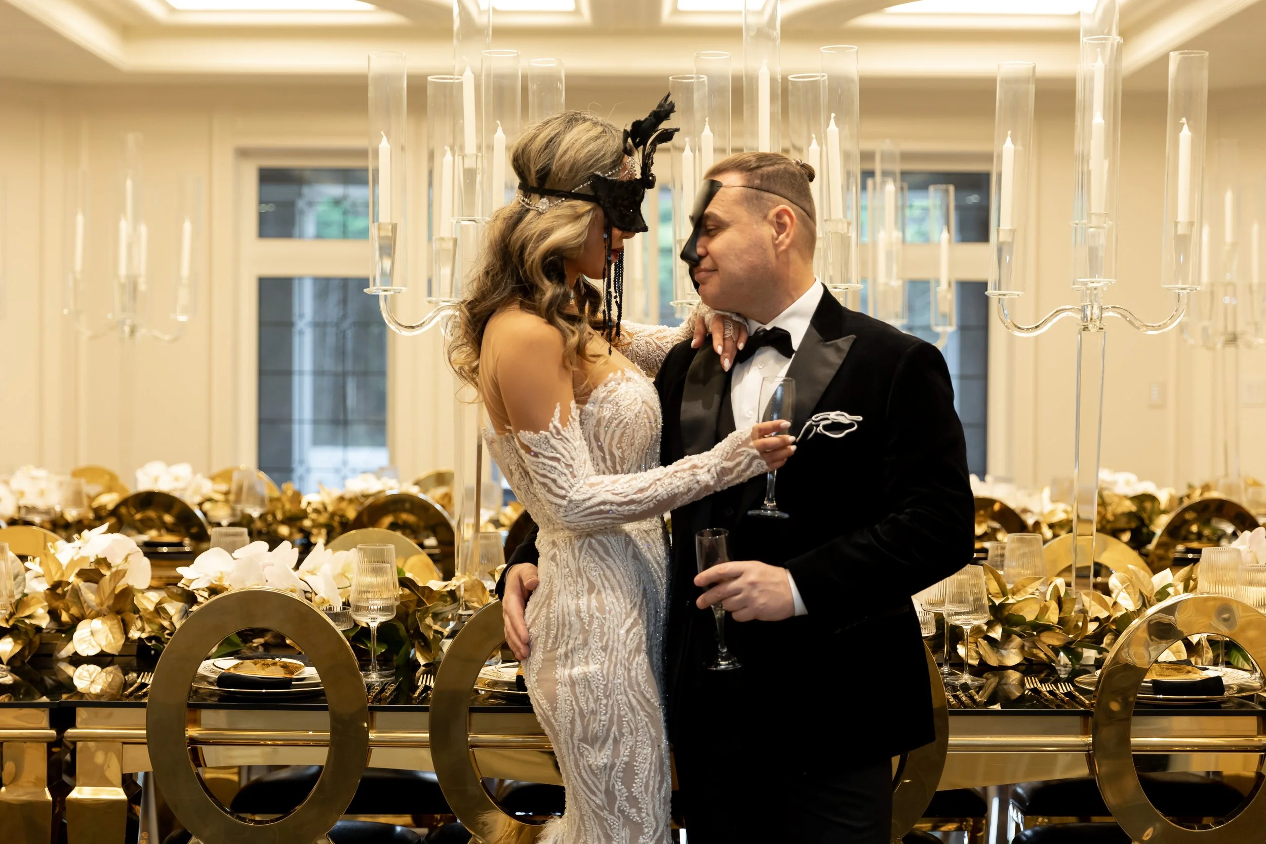 Couple dressed in formal wedding attire sharing an intimate moment, with the woman wearing a white lace wedding gown and the man in a black tuxedo, at a decorated reception table with gold accents and floral arrangements.
