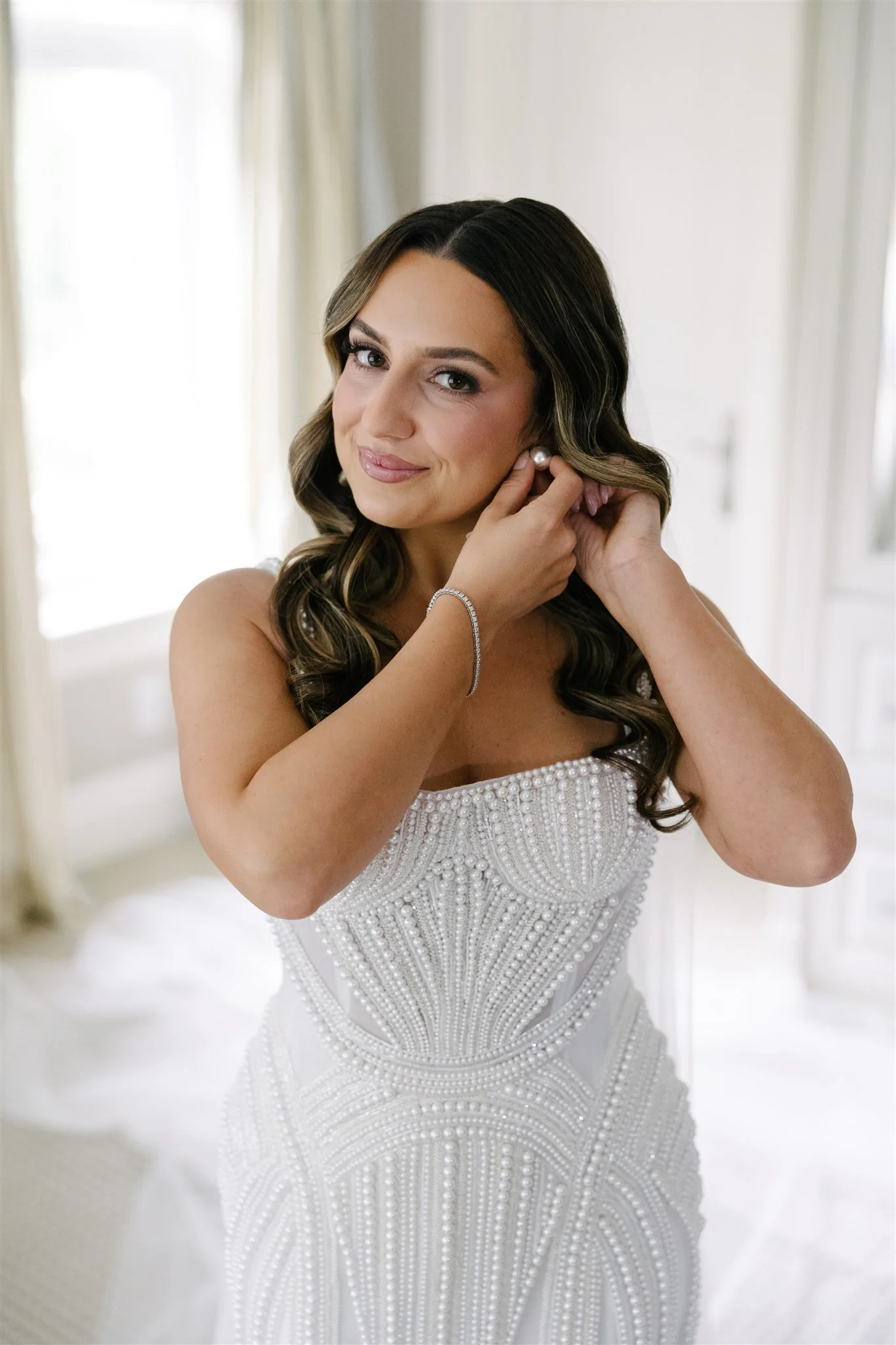 Bridal woman in a white dress with pearl embellishments, putting on pearl earrings indoors with curtains in the background.
