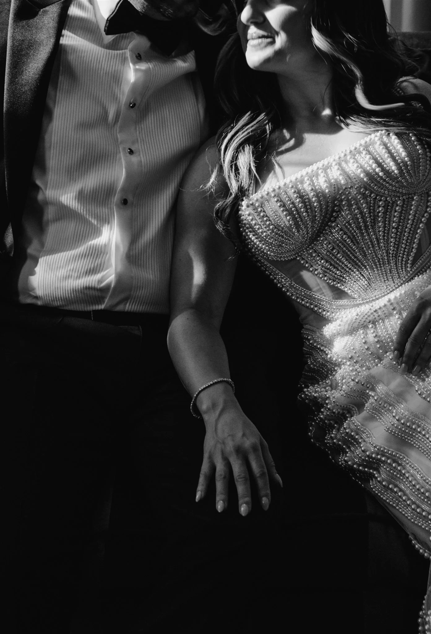 Black and white photo of a couple dressed formally, with the woman in an embellished gown and the man in a tuxedo, sitting close together.