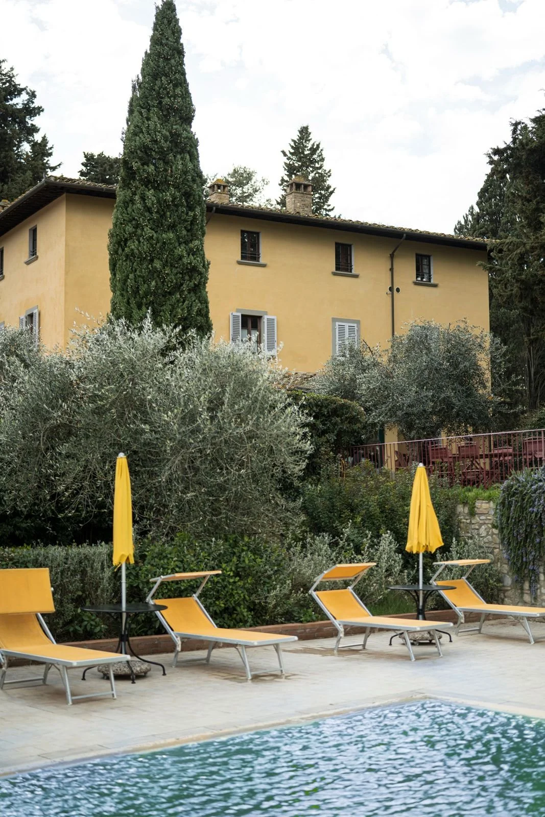 A backyard poolside scene with yellow lounge chairs and closed yellow umbrellas, surrounded by lush greenery and a yellow house in the background.
