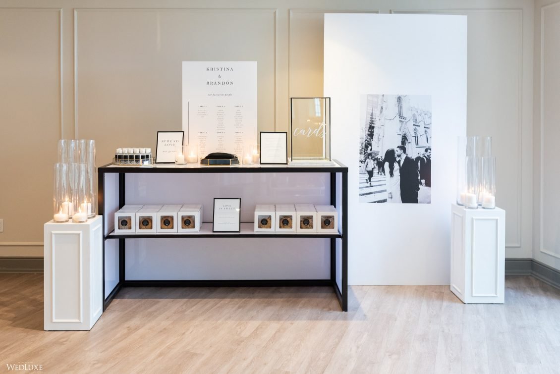 Wedding reception display with a black and white themed setup. There are candles in tall glass holders on white pedestals on either side of the arrangement. A black and white photograph of a couple in wedding attire is on the right side. The back wal