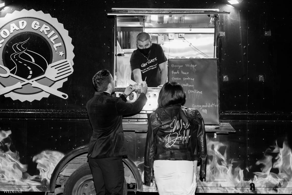 Black and white photo of a food truck named 'Road Grill' with a logo of a road and a fork and knife. Two customers, a man and a woman, are placing an order at the counter where a worker is serving food. The woman has 'Mrs. Biddle' written on the back