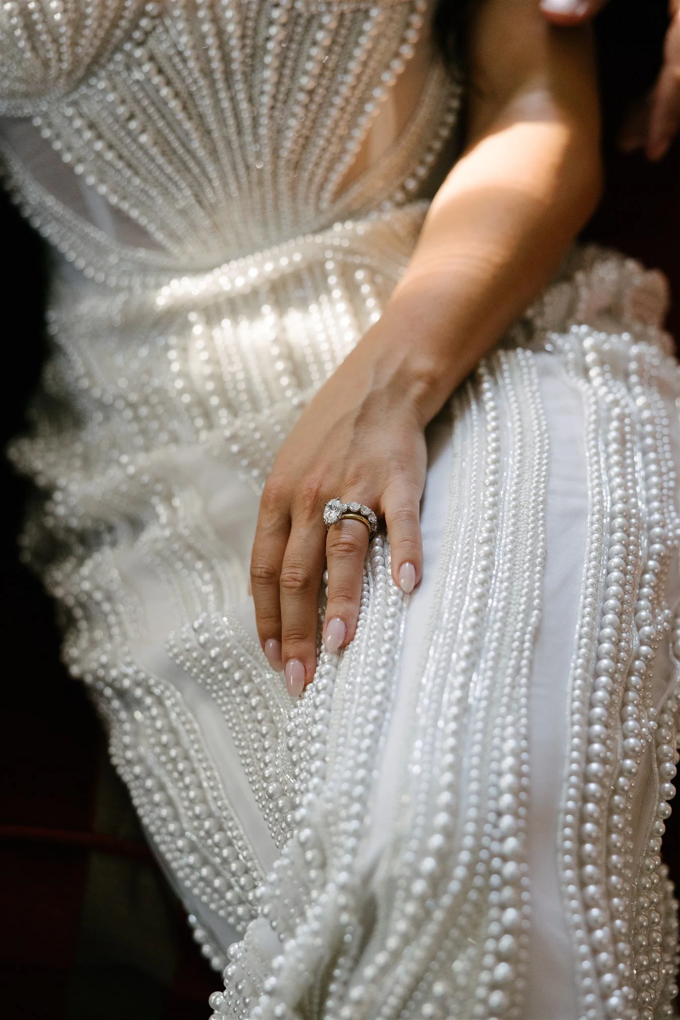 Close-up of a woman's hand resting on a white dress decorated with pearls, wearing a large diamond engagement ring.