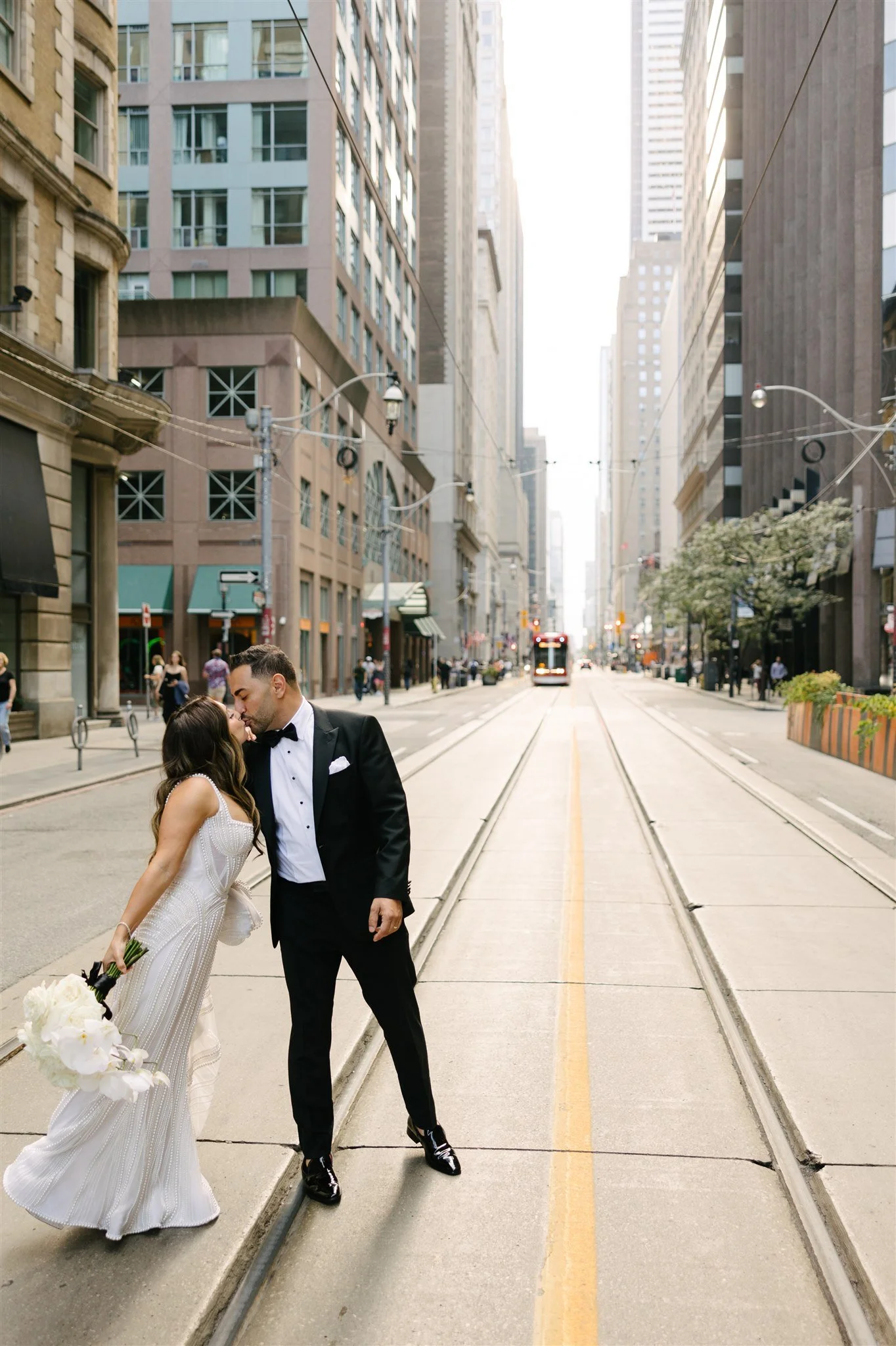 A couple in wedding attire sharing a kiss on a city street with tall buildings, streetcar tracks, and a tram in the background.