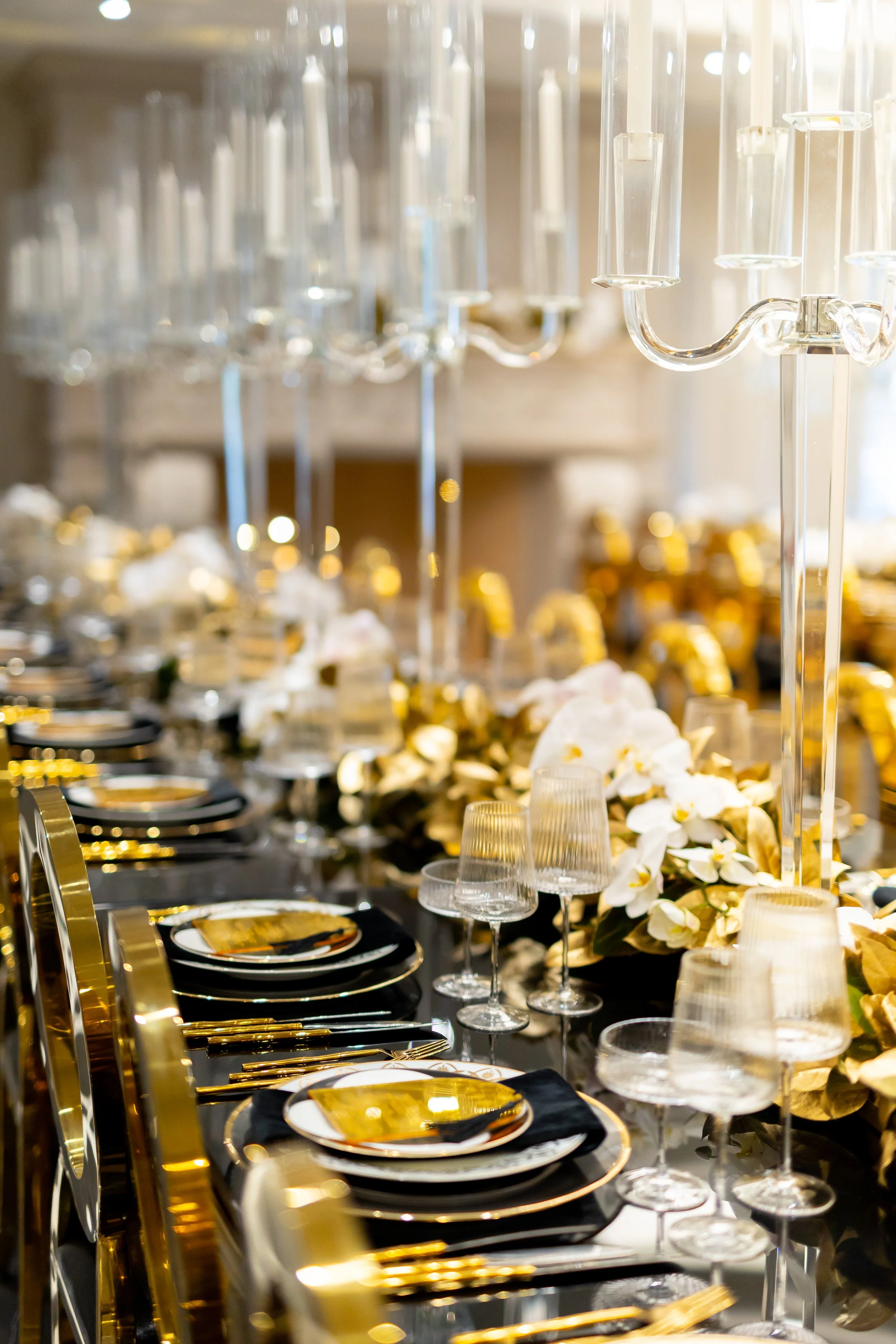 Elegant dining table set with gold accents, black napkins, white orchids, and tall glass candle holders, prepared for a formal event.