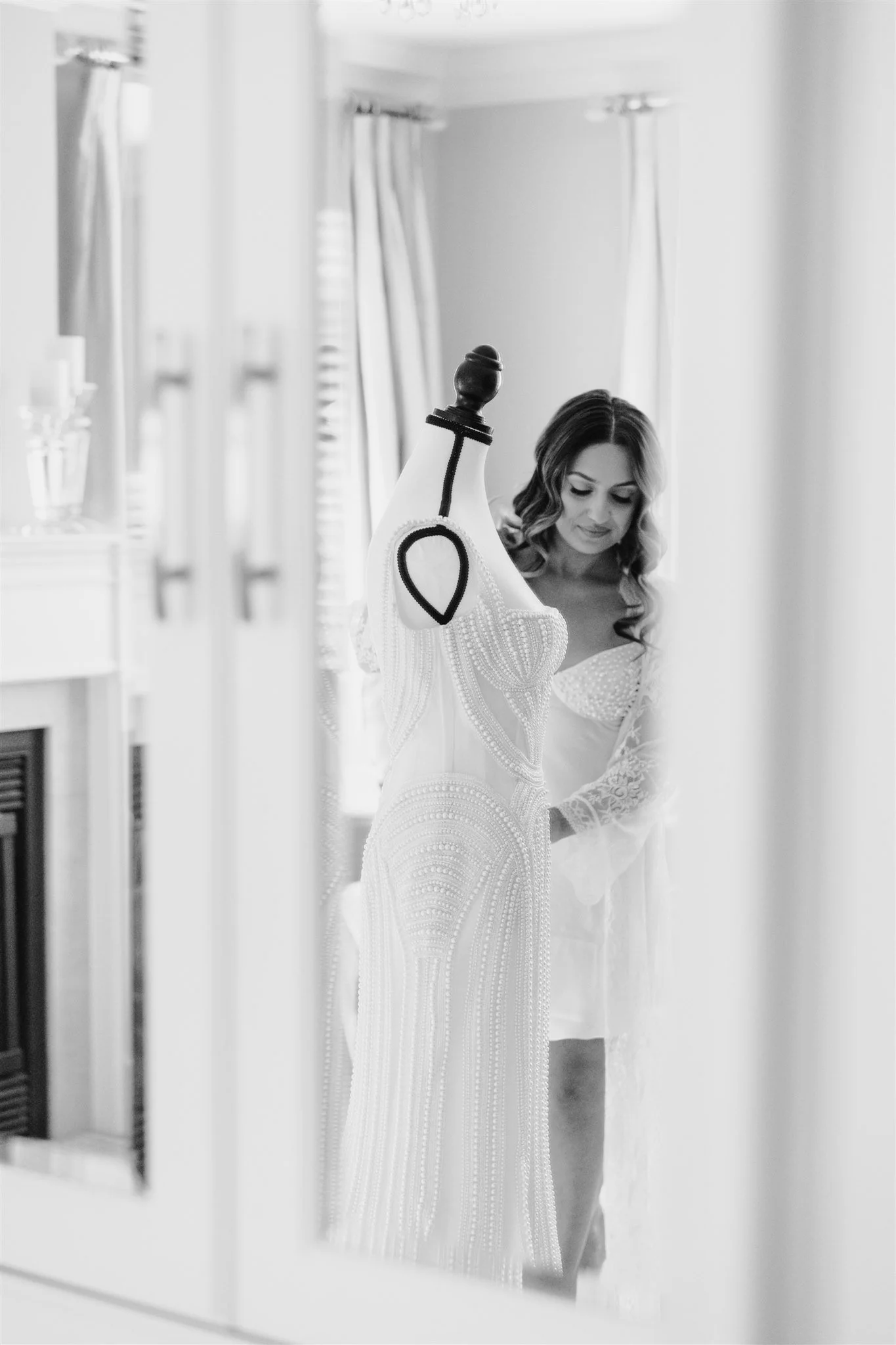 A woman is looking at a wedding dress on a mannequin, reflected in a mirror in a room with light-colored curtains.