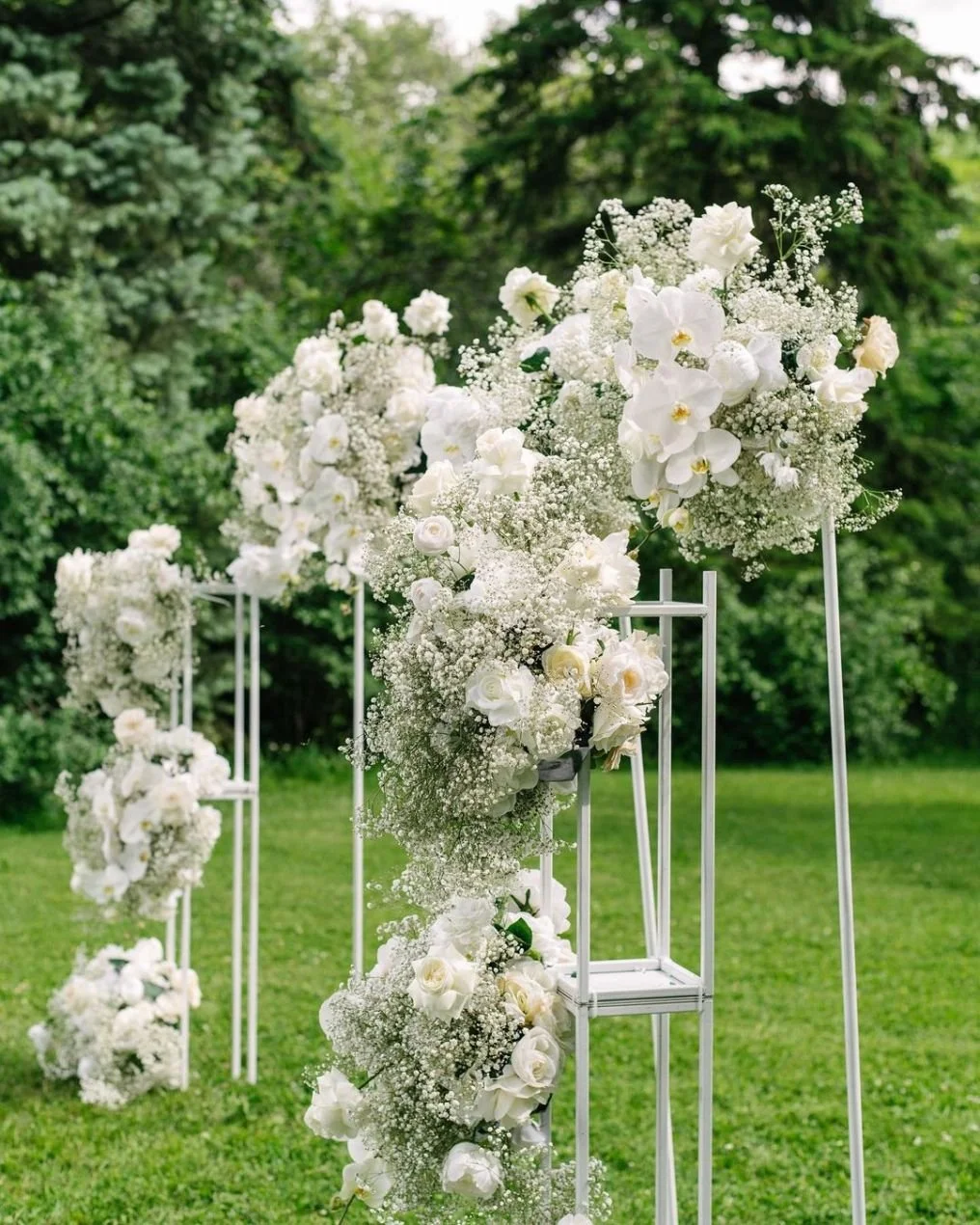White floral arrangement with roses and baby's breath on a metal stand outdoors, with green trees and grass in the background.