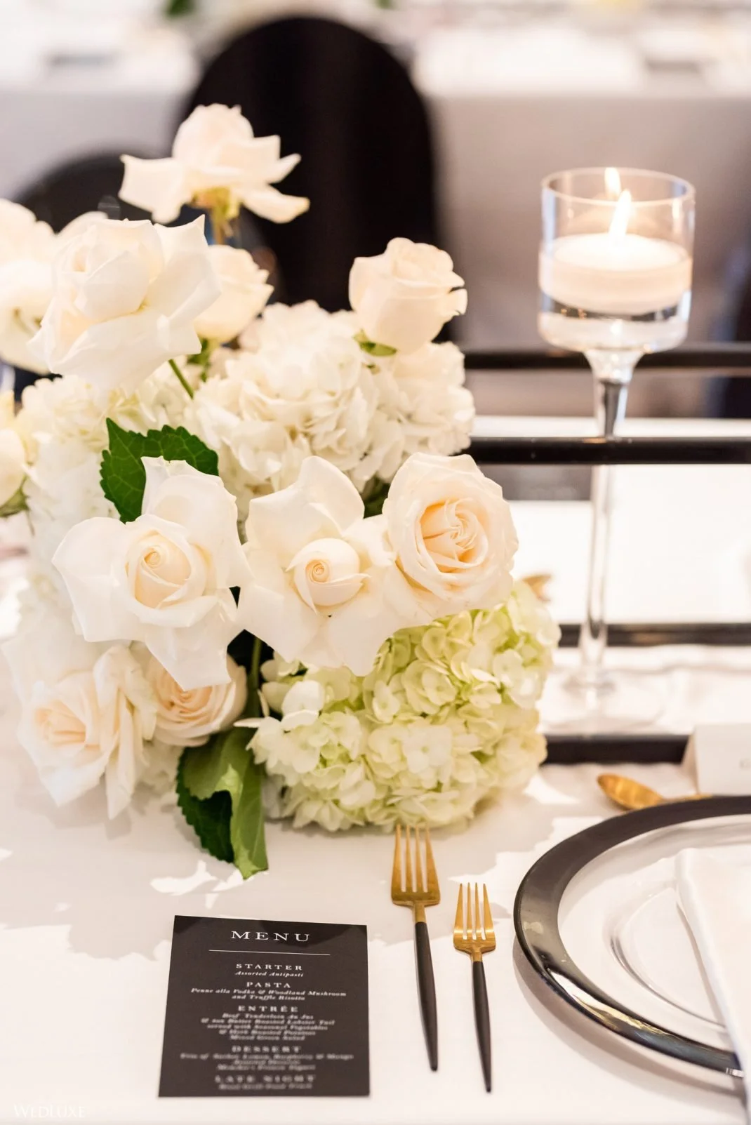A table setting with a floral centerpiece of white roses and hydrangeas, a black menu card, gold and black cutlery, a black and silver plate, and a candle in a glass holder, on a white table.