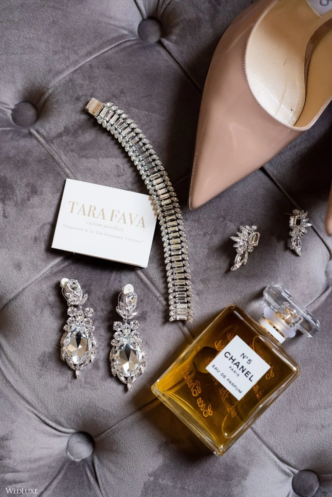 A collection of jewelry including a diamond necklace, earrings, and a business card that reads "Tara Fava" resting on a gray, tufted surface next to a beige high-heeled shoe and a bottle of Chanel No. 5 perfume.
