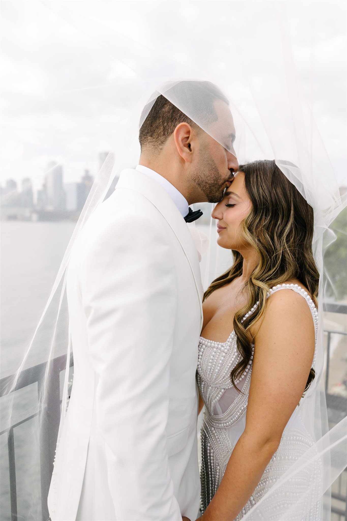 A bride and groom sharing a moment under a veil on a balcony with city skyline in the background.