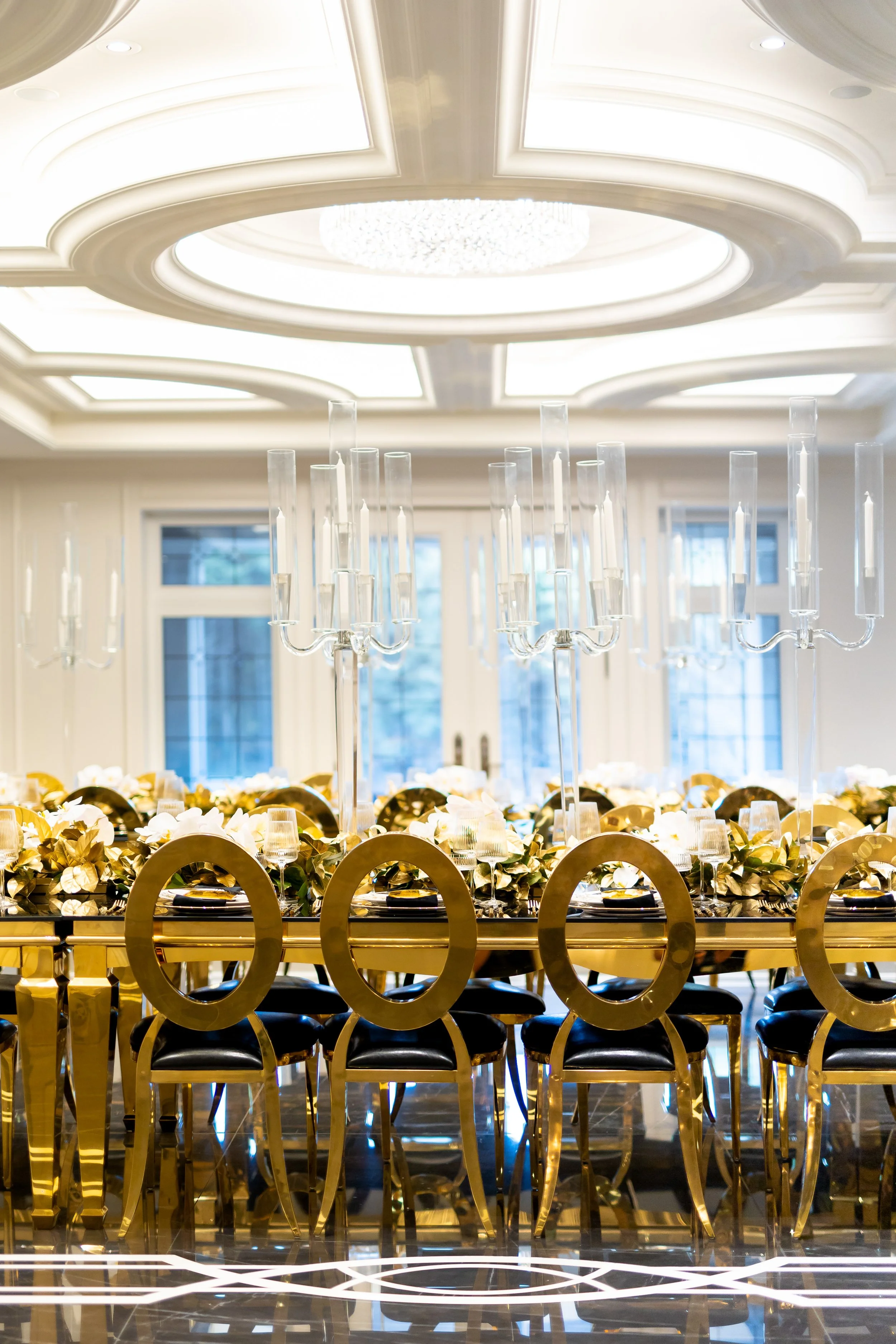 Elegant dining table with gold chairs, tall glass candleholders, and floral centerpieces in a luxurious room with a decorative ceiling and large windows.