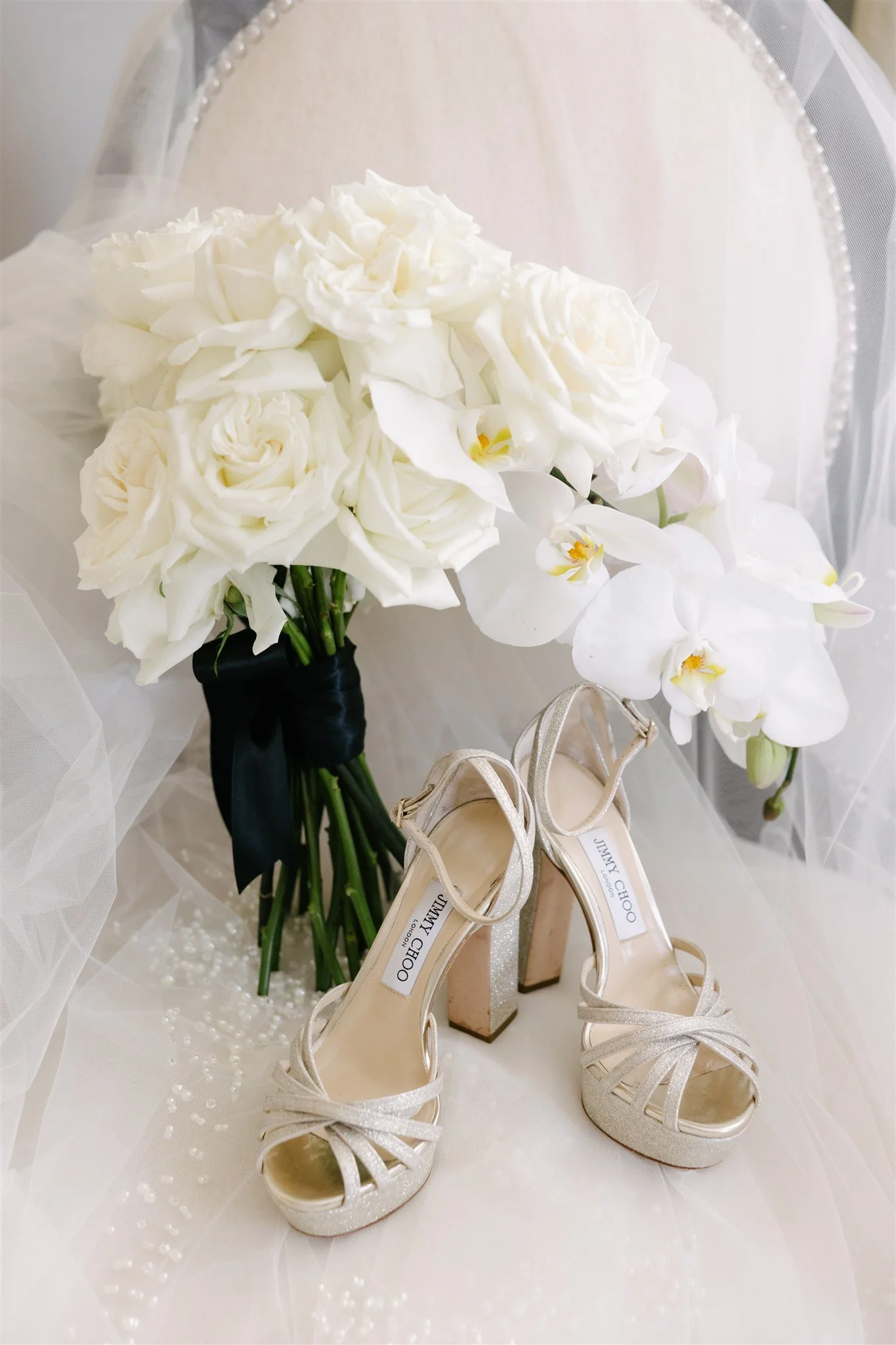 A bouquet of white roses and white orchids resting on a white surface with a pair of silver Jimmy Choo high-heeled shoes in front.
