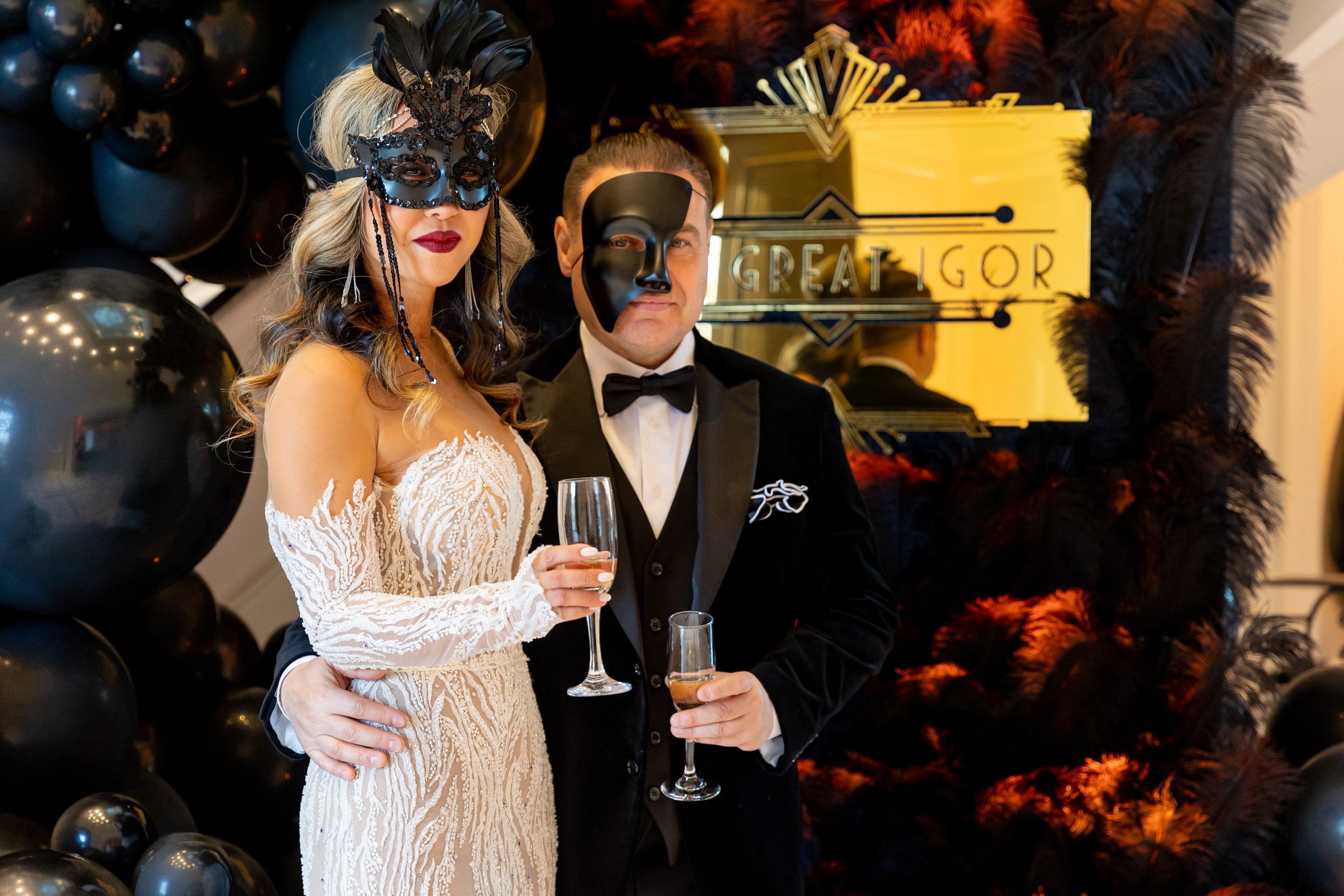 A woman in an elegant beige lace dress and a man in a black tuxedo with a mask are at a party, holding champagne glasses, with black balloons and a decorative black and red feathered backdrop.