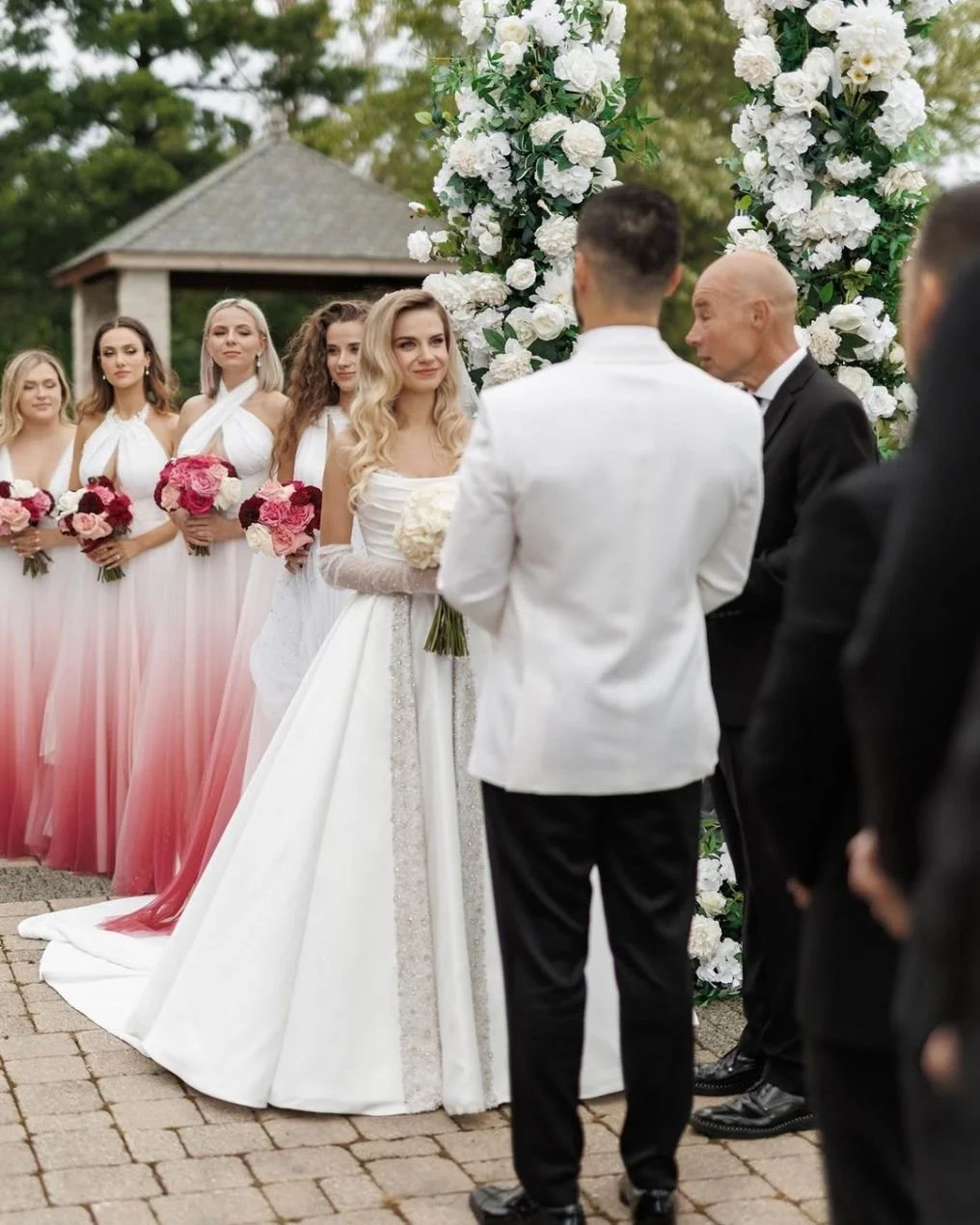 A wedding ceremony with a bride in a white dress holding a bouquet, groom in a white tuxedo, officiant, and bridesmaids in white and pink dresses holding pink flowers, outdoors near a floral arch.